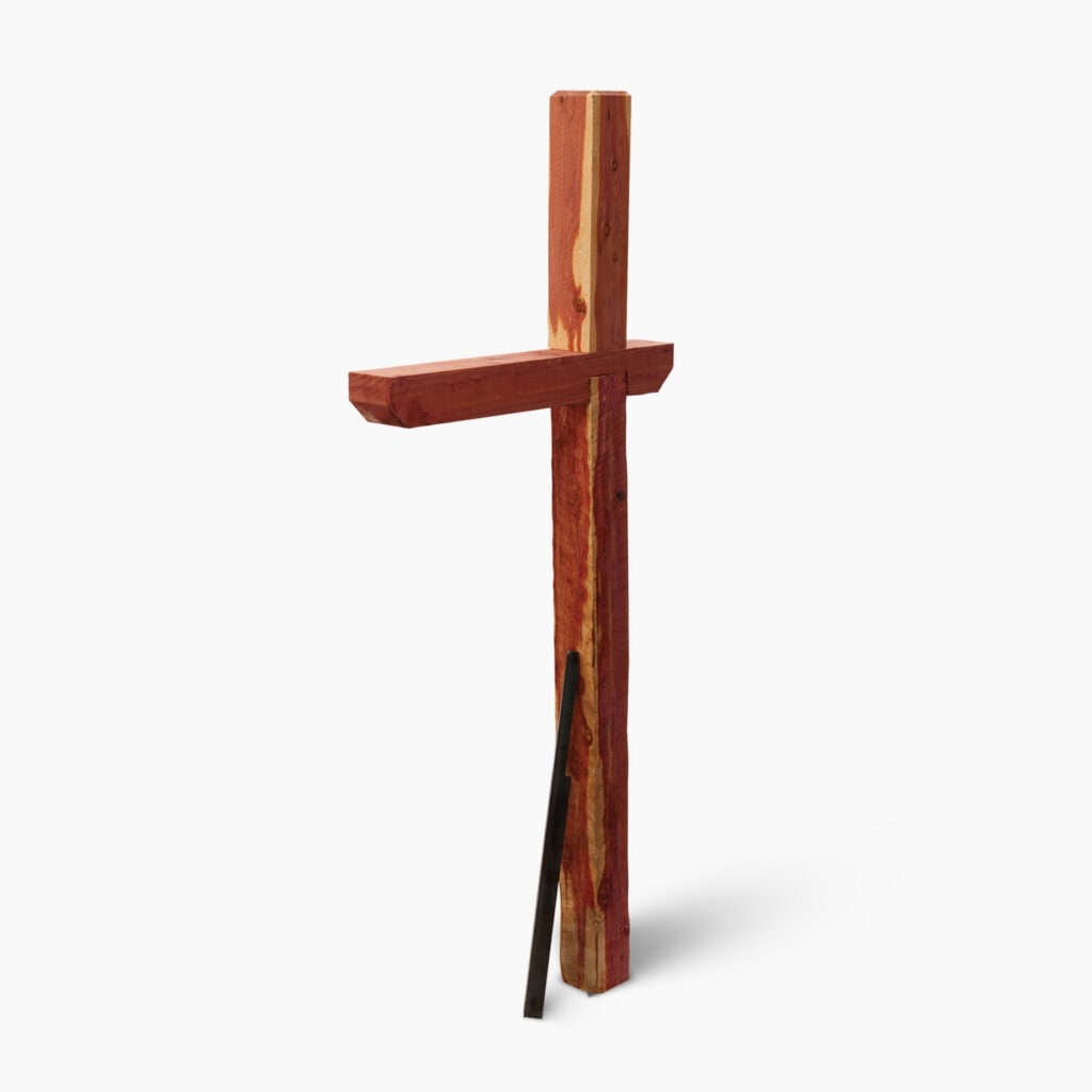 Rough wooden cross painted reddish-brown with a vertical post and horizontal beam on a white background; a dark stake rests at the base.