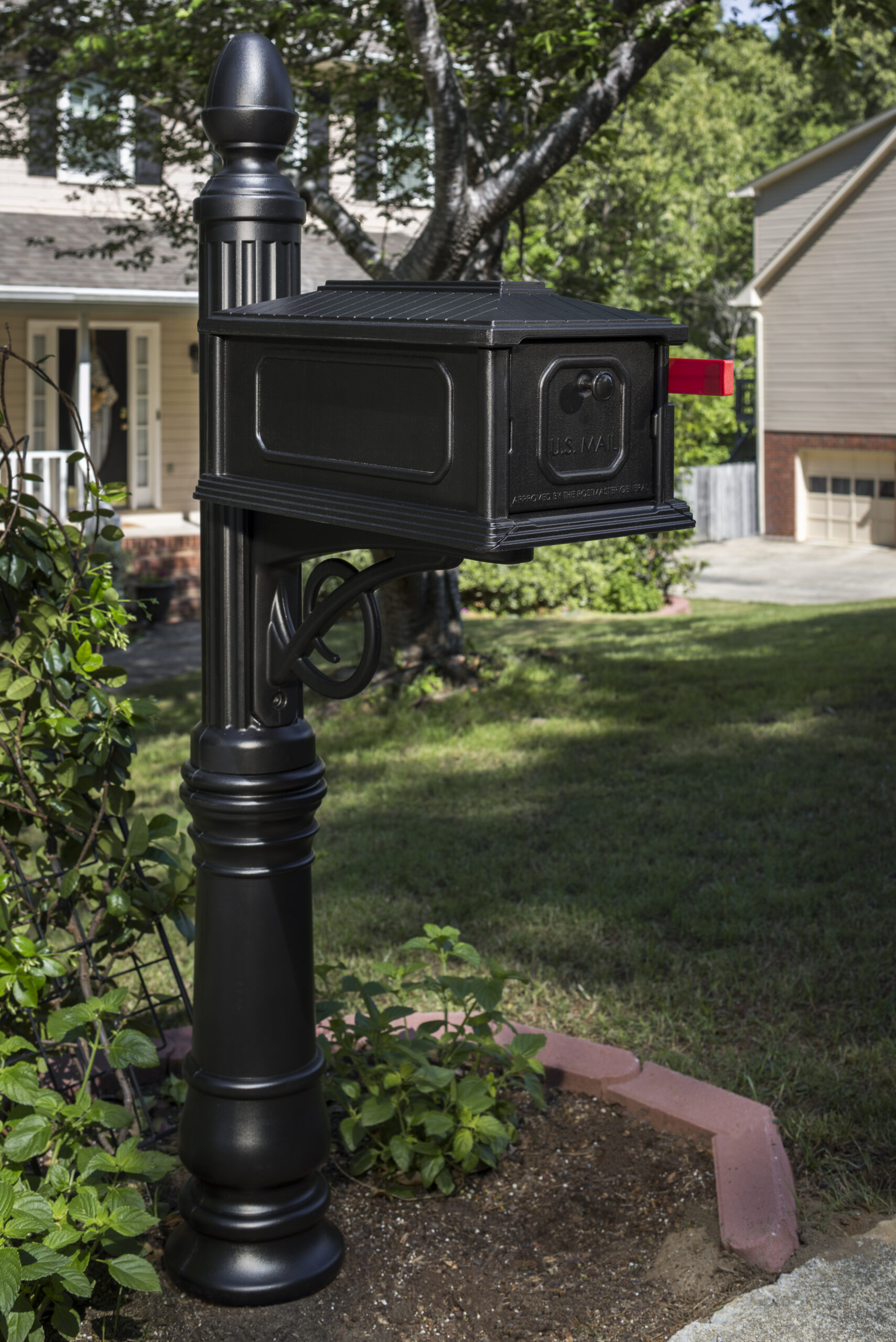 Black U.S. Mail mailbox mounted on an ornate black post in a suburban front yard, red flag raised on the side edge.