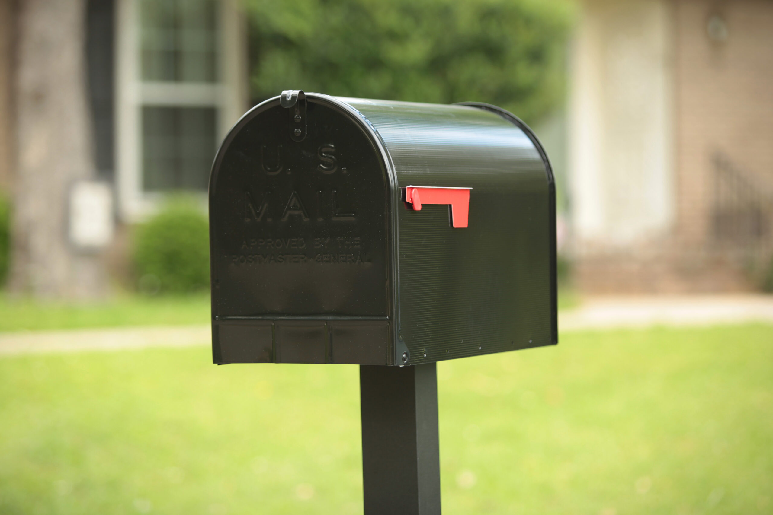 Black US mailbox with red flag raised on a post in a sunny front yard.
