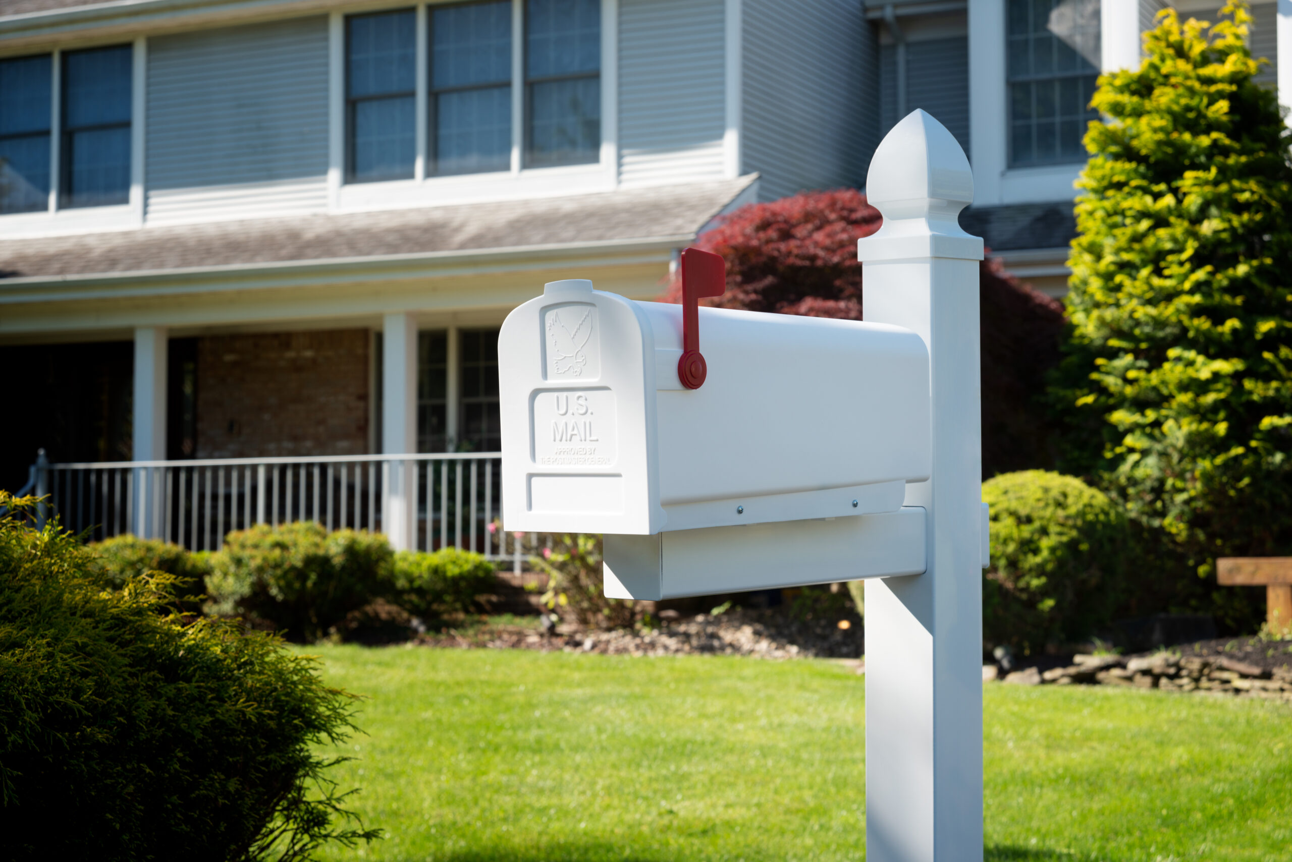 White U.S. mailbox on a post in a front yard, red flag up, in front of a suburban house with a green lawn.