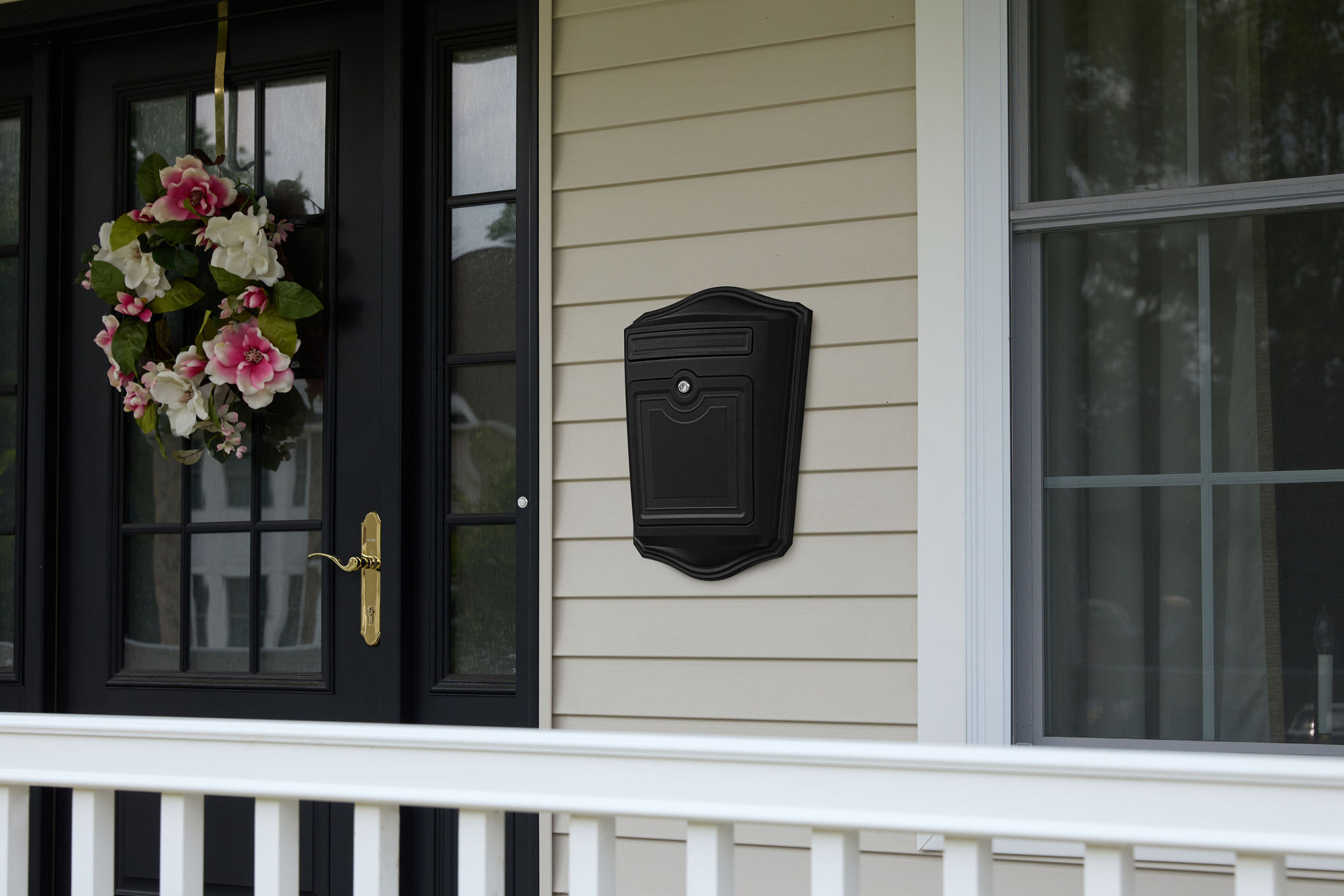 Black front entry with gold handle, pink-white flower wreath on the left door, beige siding, and a wall-mounted black mailbox on the wall beside it.
