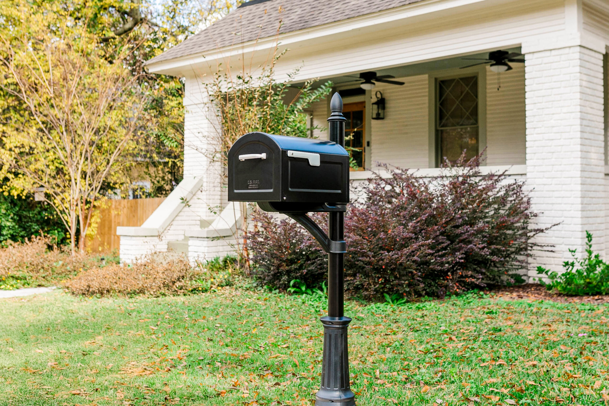 Black mailbox mounted on a decorative post in a green front yard with a white brick house in the background