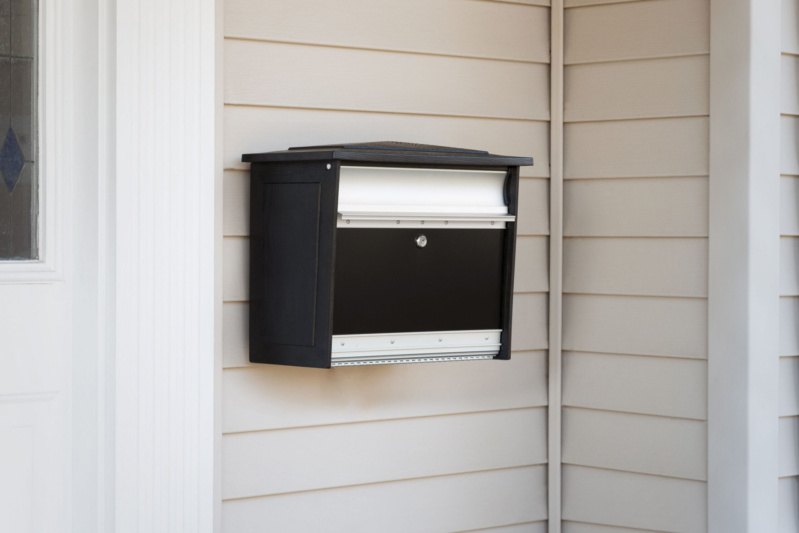 A black exterior wall mailbox mounted to beige siding with a horizontal slot and a small round lock in the center