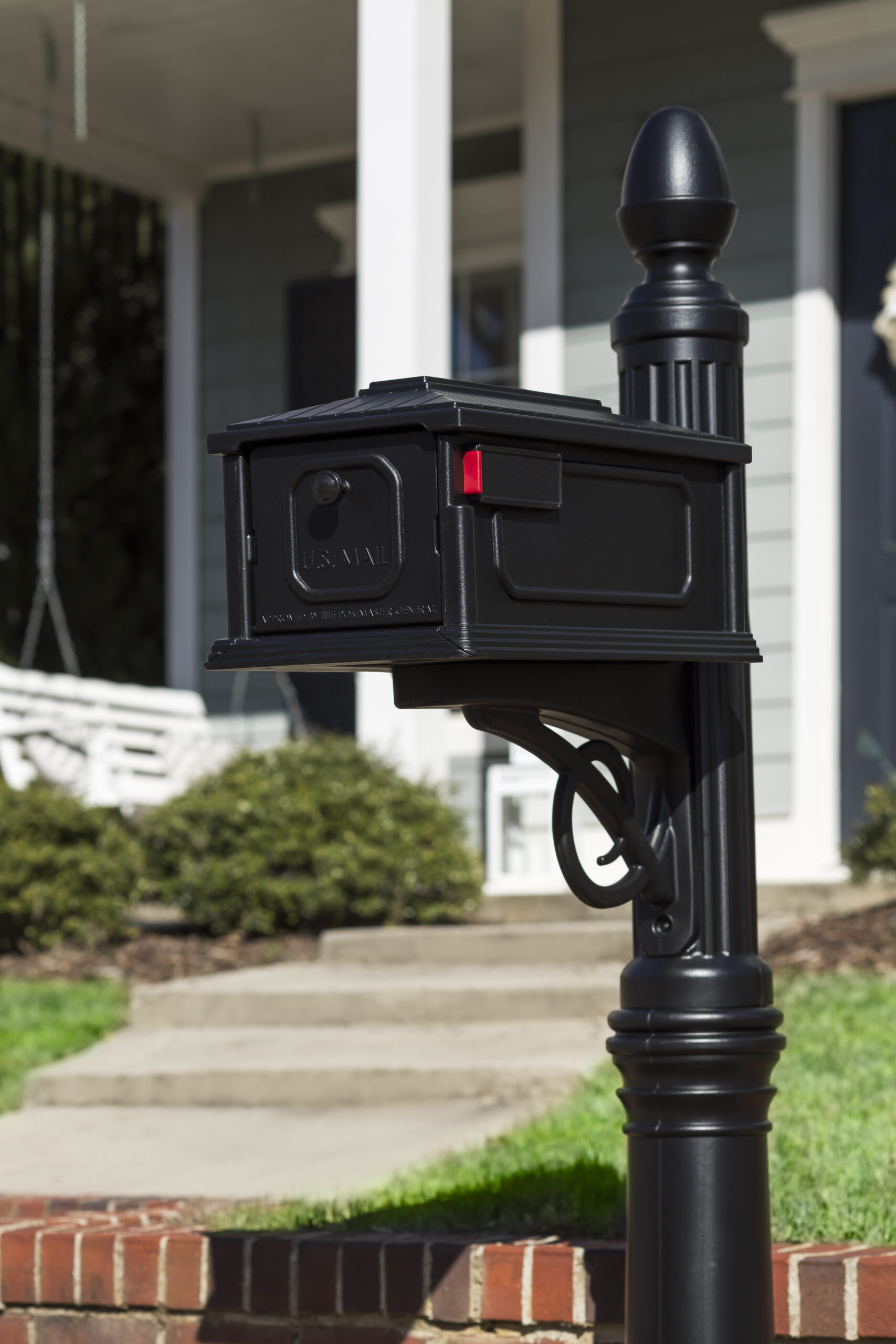 Black metal mailbox mounted on a tall decorative post in a suburban front yard with steps and shrubbery in the background, near a brick path.