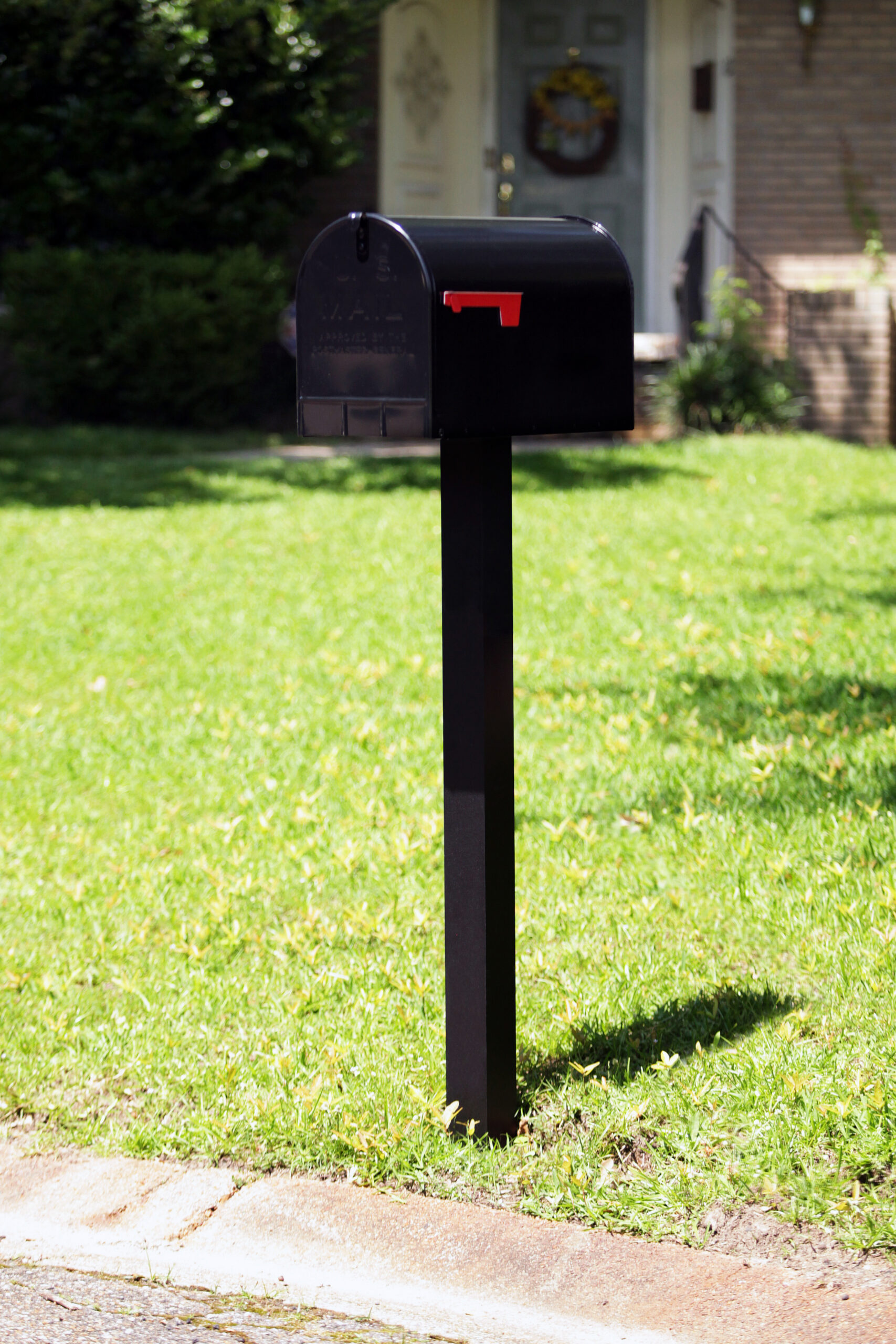 Black mailbox with red flag mounted on a tall post in a green front yard of a suburban home at the curbside edge.