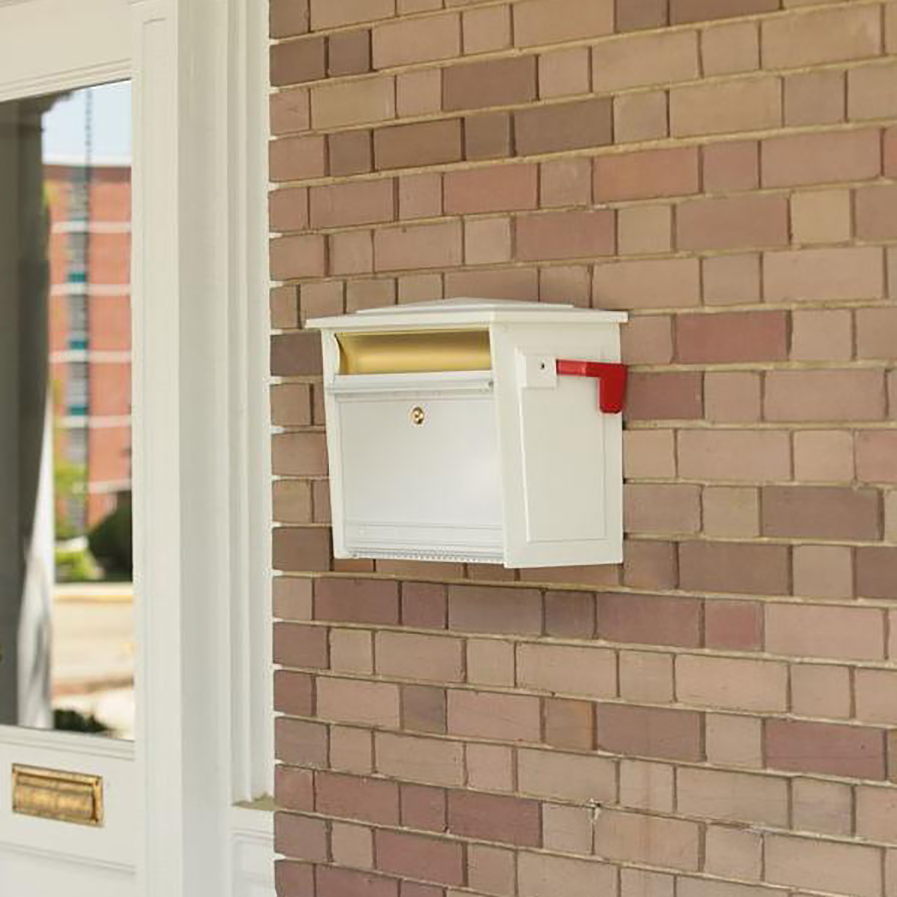 White wall-mounted mailbox with a gold flag on a brick exterior near a white door entrance, ready for mail pickup