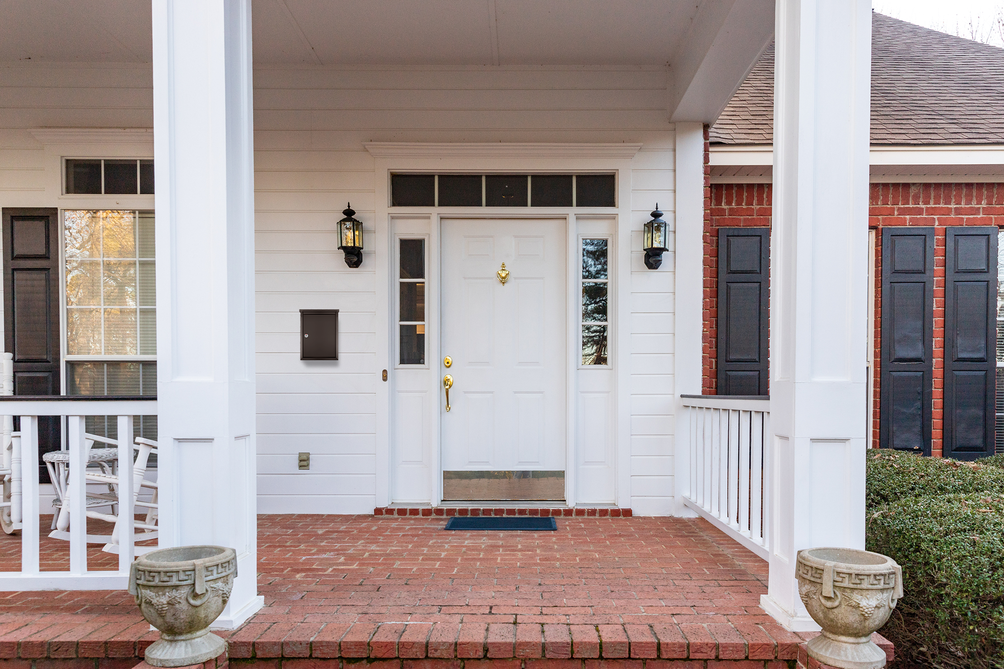 White front entryway with a paneled door, sidelights, brass hardware, and wall lanterns on white siding, brick porch, and matching columns.