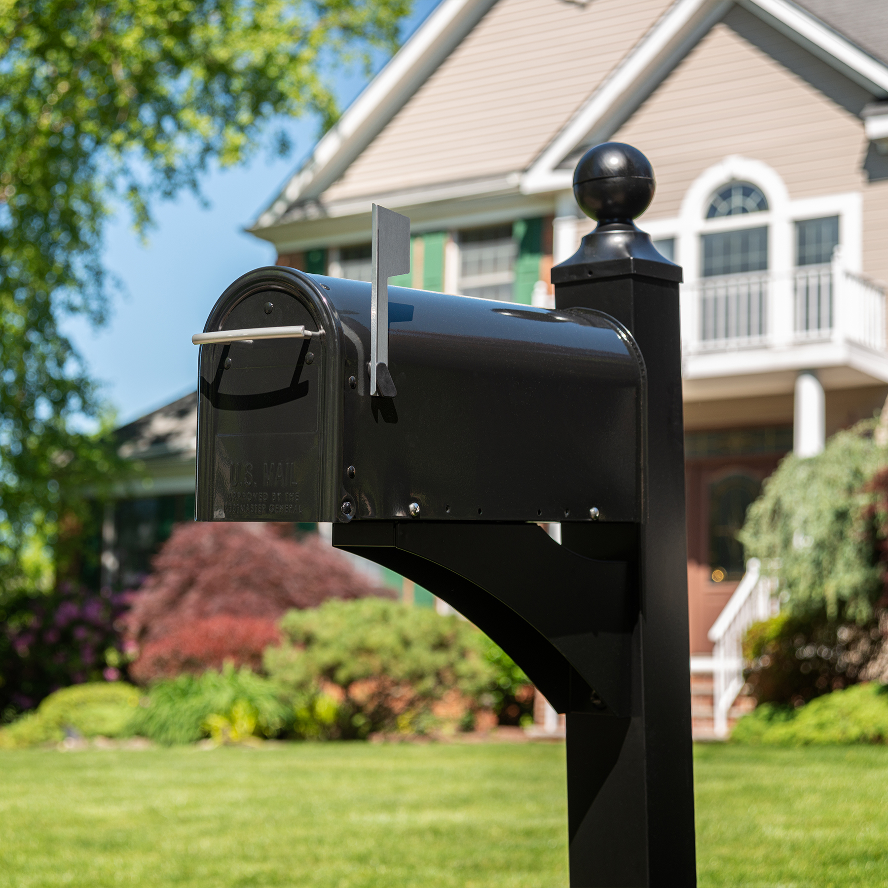 Black metal mailbox on a post in a sunny suburban front yard with a beige house and green lawn.
