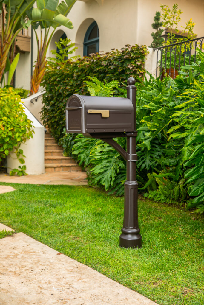Dark brown curbside mailbox mounted on a decorative post in a landscaped yard, with a beige house and lush green plants in the background.