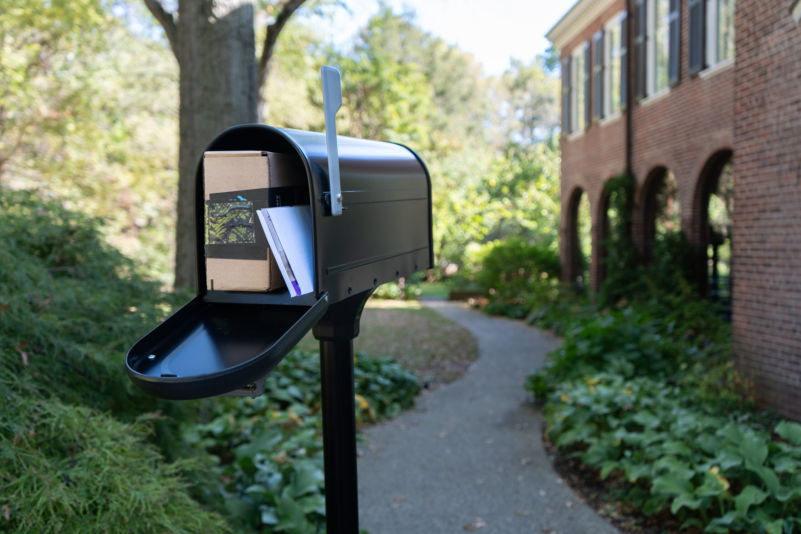Open mailbox on a post with letters and a small package visible, along a sidewalk beside a brick building and greenery.