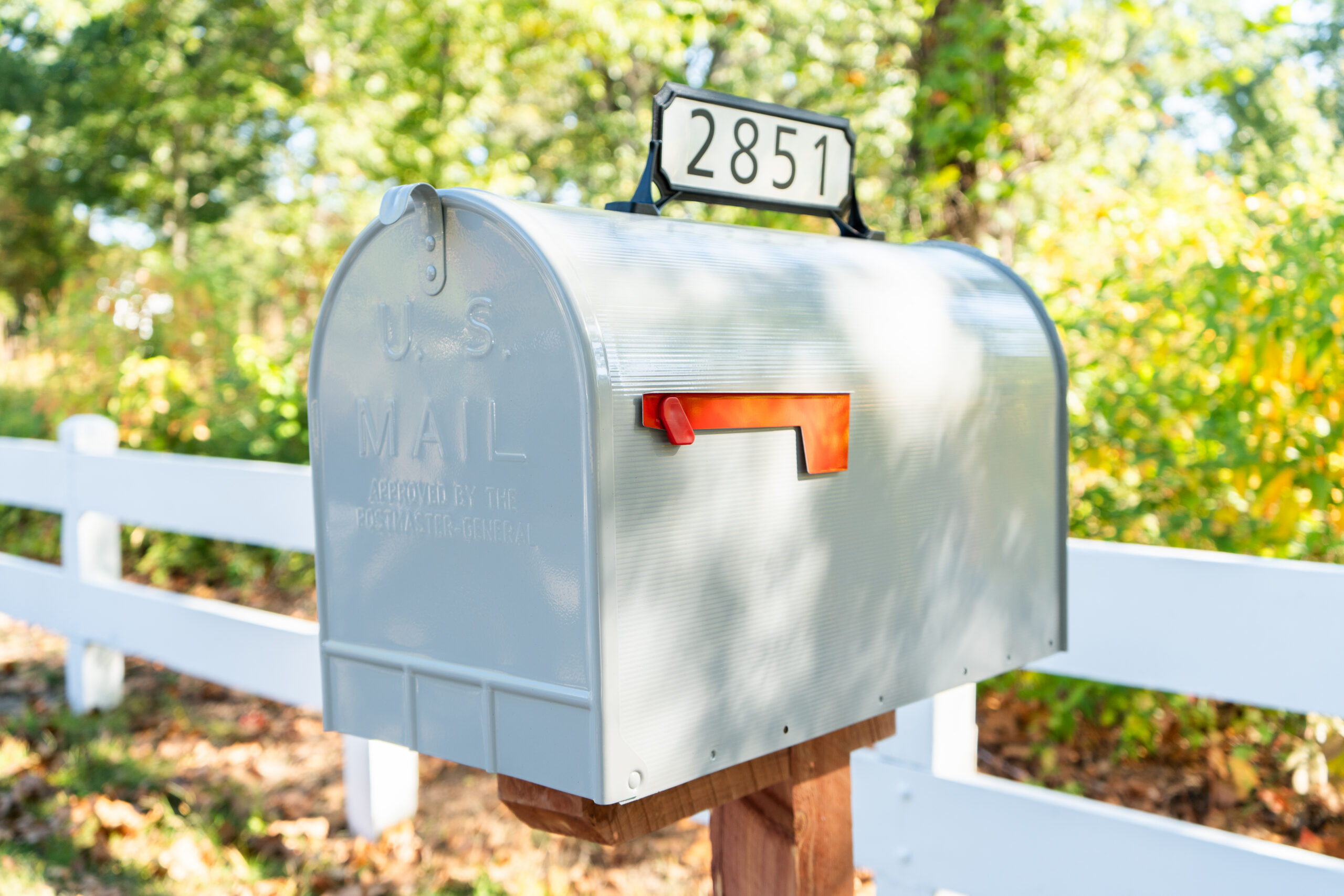 Silver US mail mailbox with red flag raised, mounted on a wooden post beside a white fence in a sunny yard.