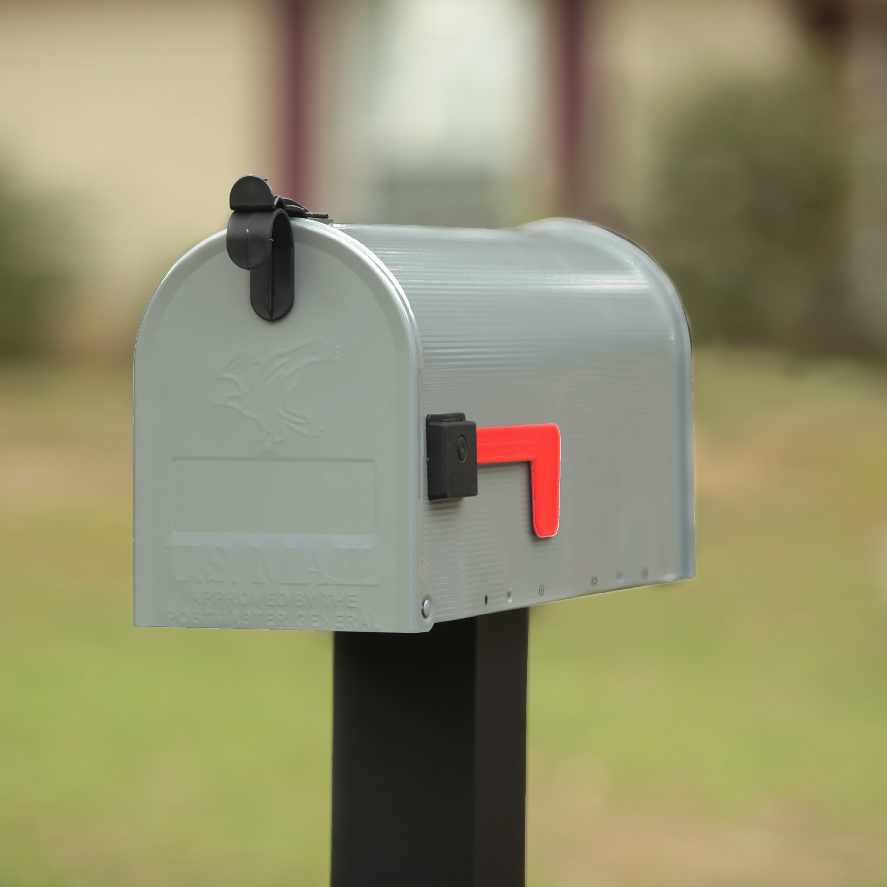 Gray metal curbside mailbox with its red flag raised, mounted on a black post in a yard.