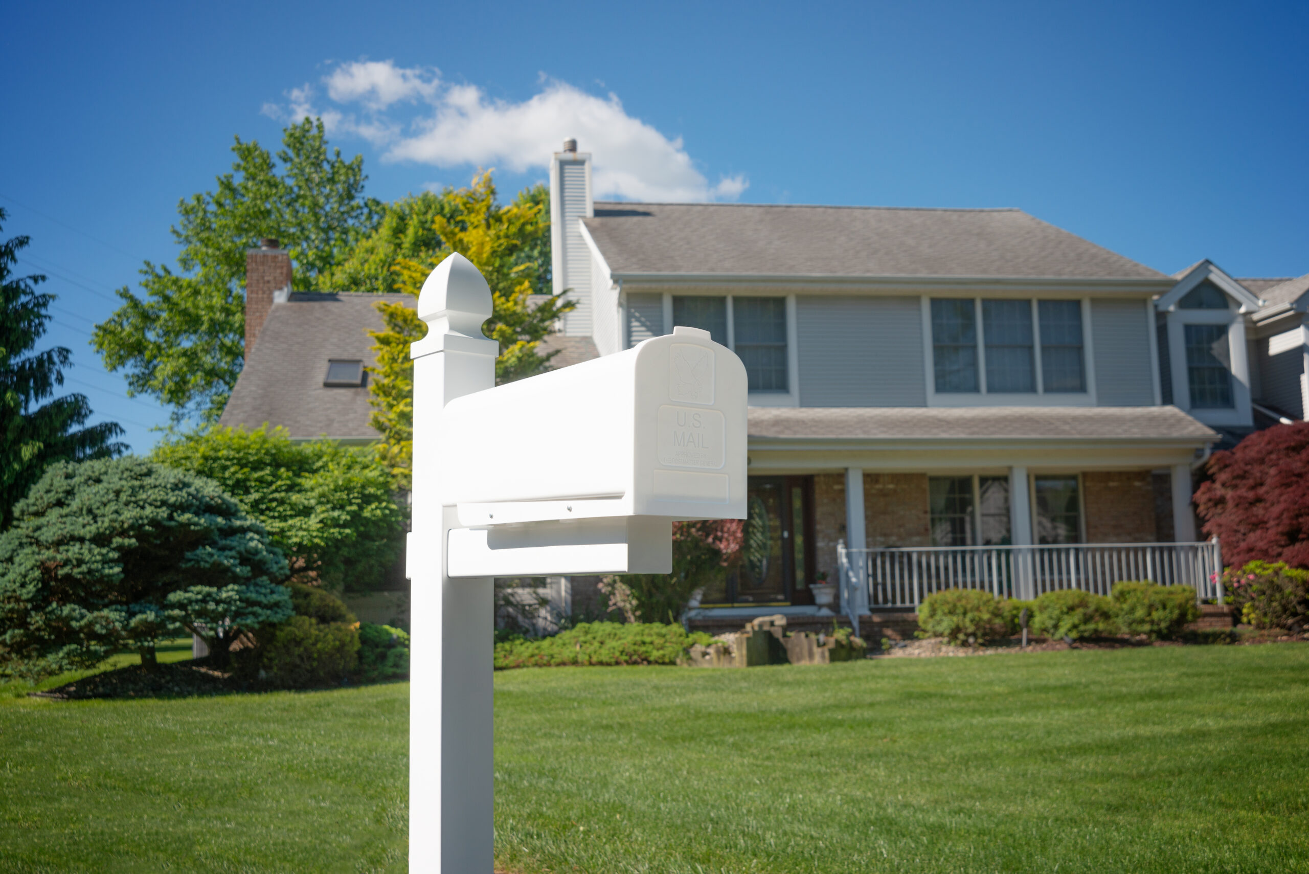 White mailbox on a post in a front-yard with a two-story suburban house and a green lawn in the background.