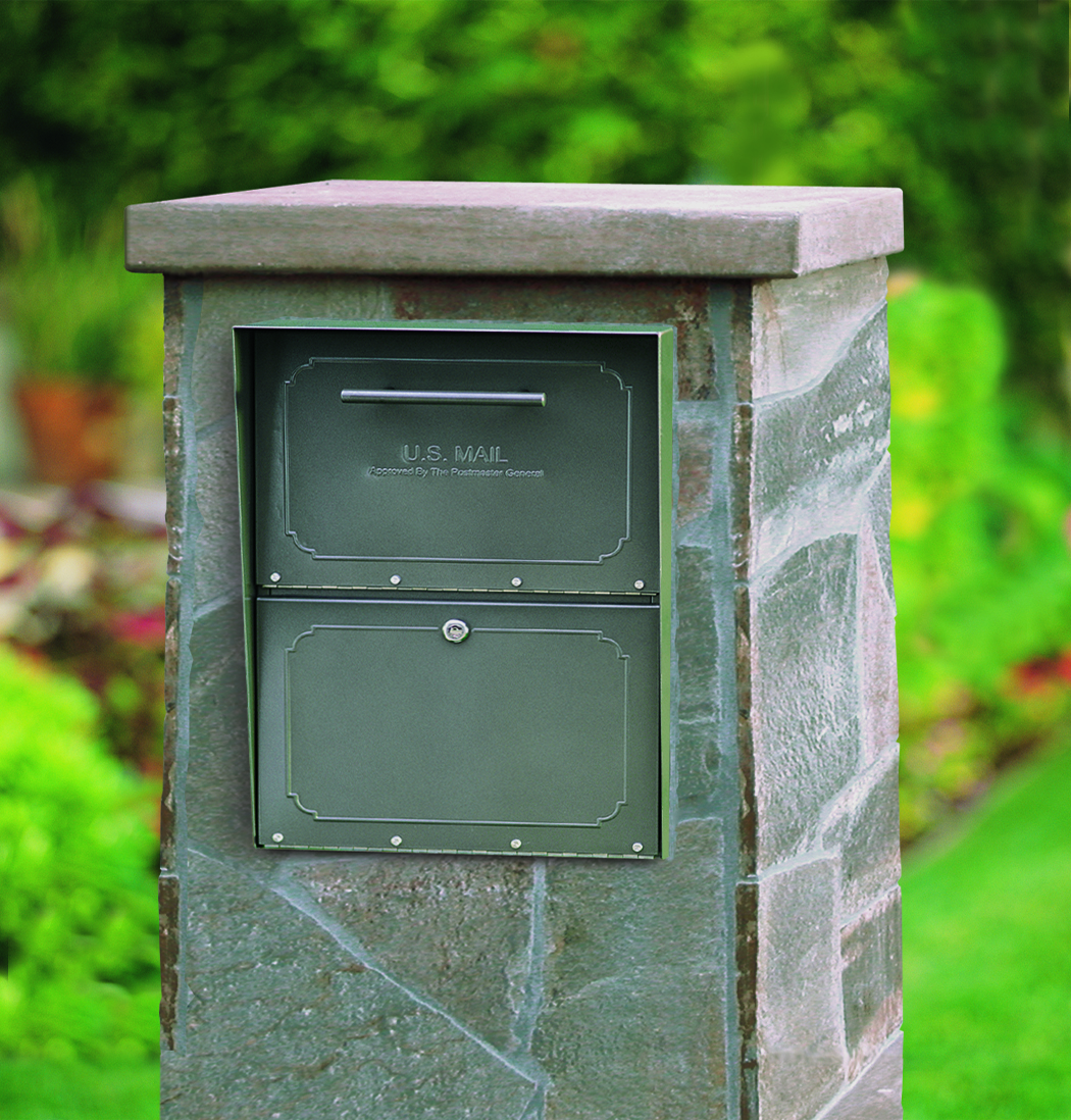 Green U.S. Mailbox with two compartments mounted on a stone pedestal outdoors in a garden setting, ready for mail delivery.