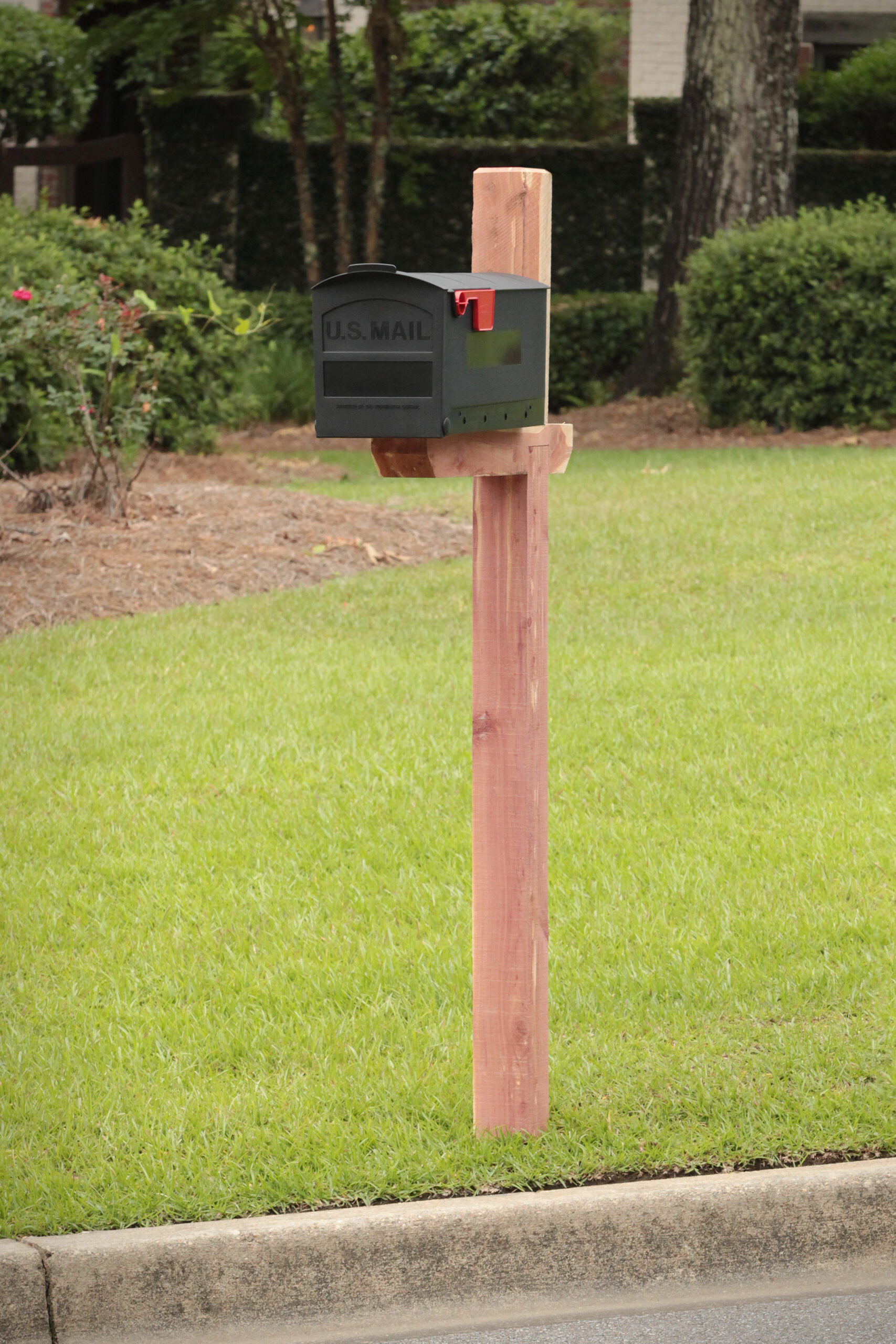 Dark gray U.S. Mailbox with red flag raised, mounted on a wooden post in a green suburban front yard.