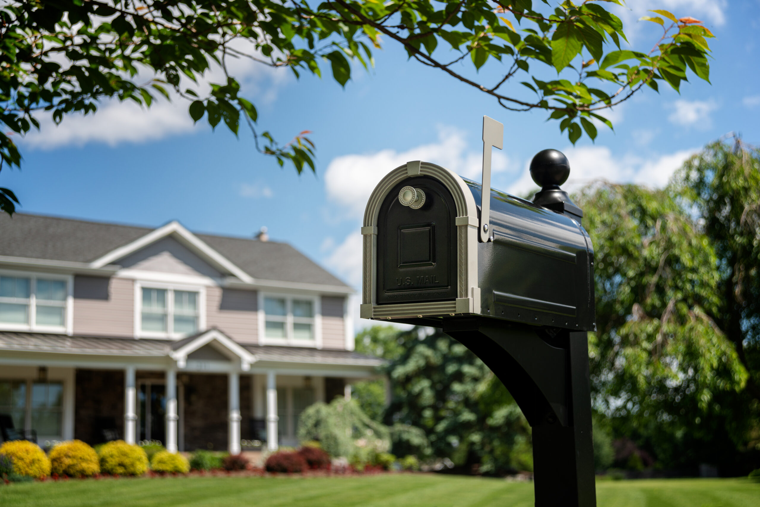 Black mailbox on a post in a front yard; beige two-story house with white trim and trimmed shrubs in the background.