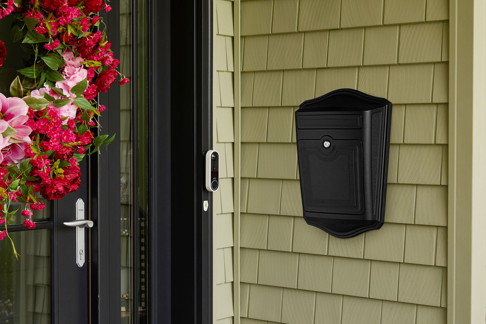 Black wall-mounted mailbox on beige siding beside a glass front door, with a pink-red flower arrangement hanging on the left.