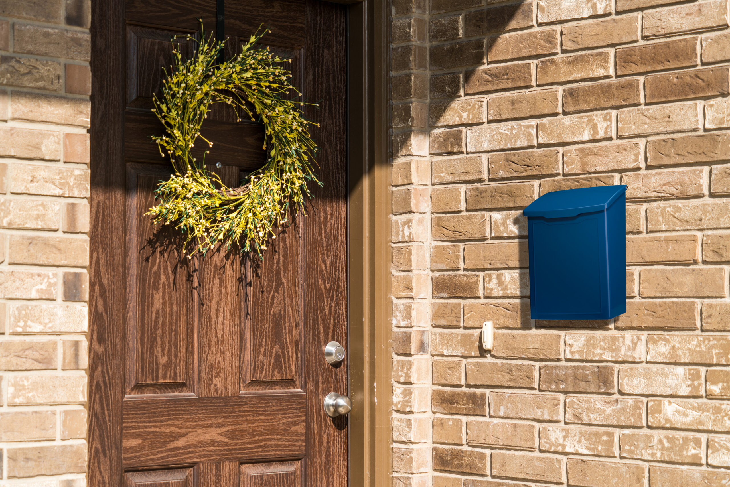 Wooden front door with a green wreath, next to a blue wall mailbox on a beige brick exterior.
