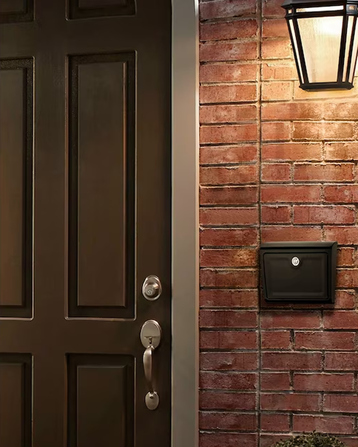 Front entryway with a dark brown paneled door, silver doorknob and deadbolt, brick wall, wall mailbox, and a lit lantern above.