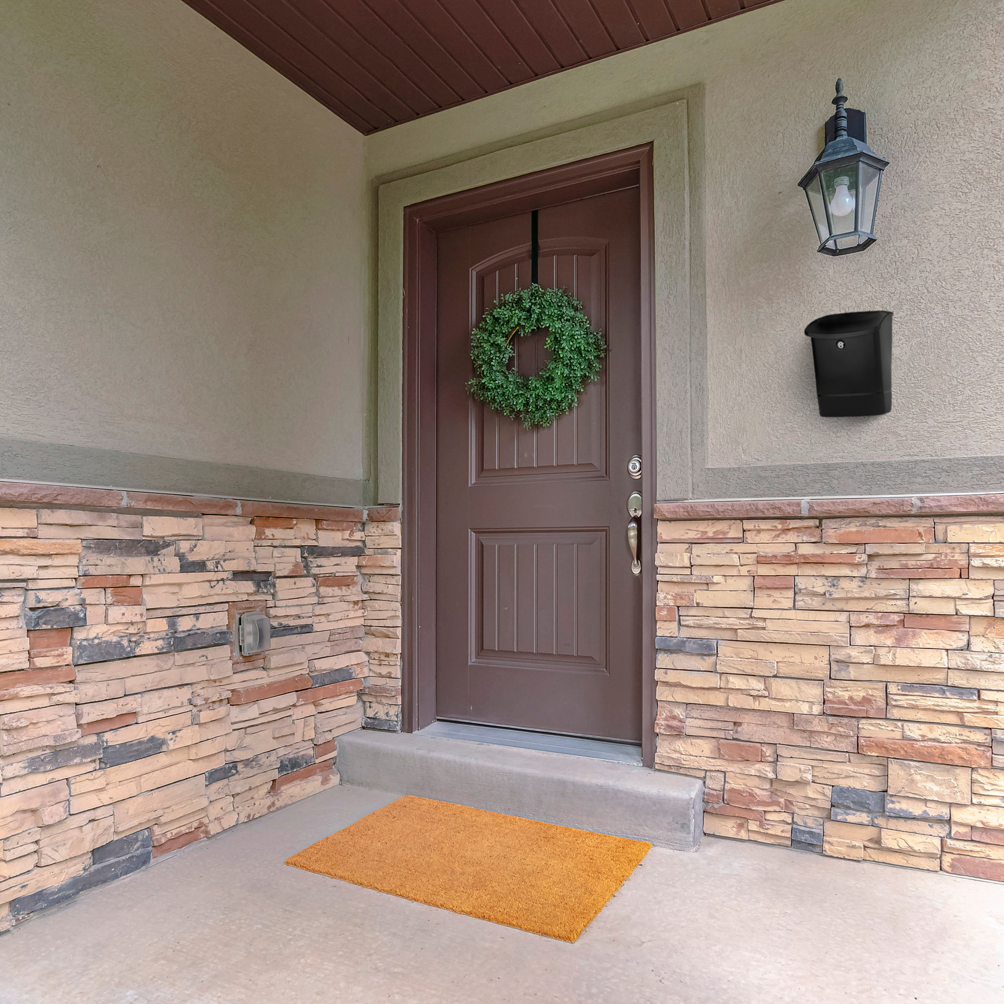 Brown front door with a green wreath, beige stucco, and stacked-stone lower wall; orange doormat at the entry with an exterior lantern nearby.