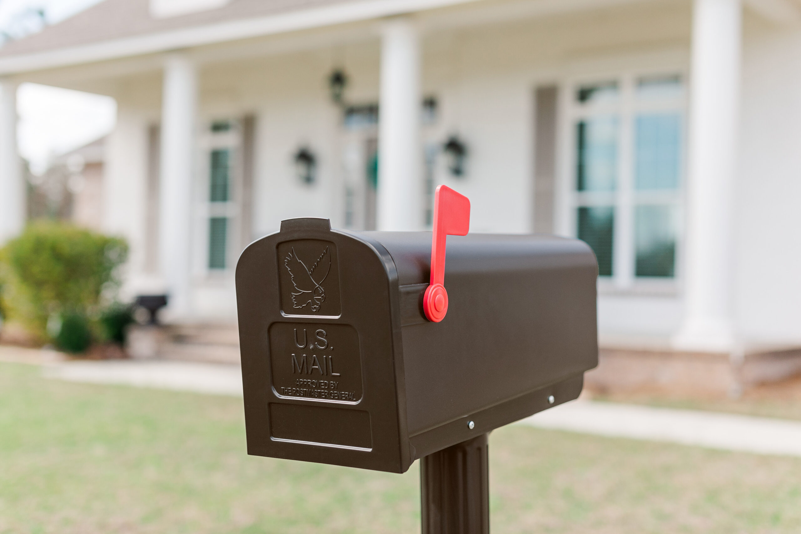 Black U.S. Mail mailbox with red flag raised in a suburban yard, mailbox near a white house with columns in the background.