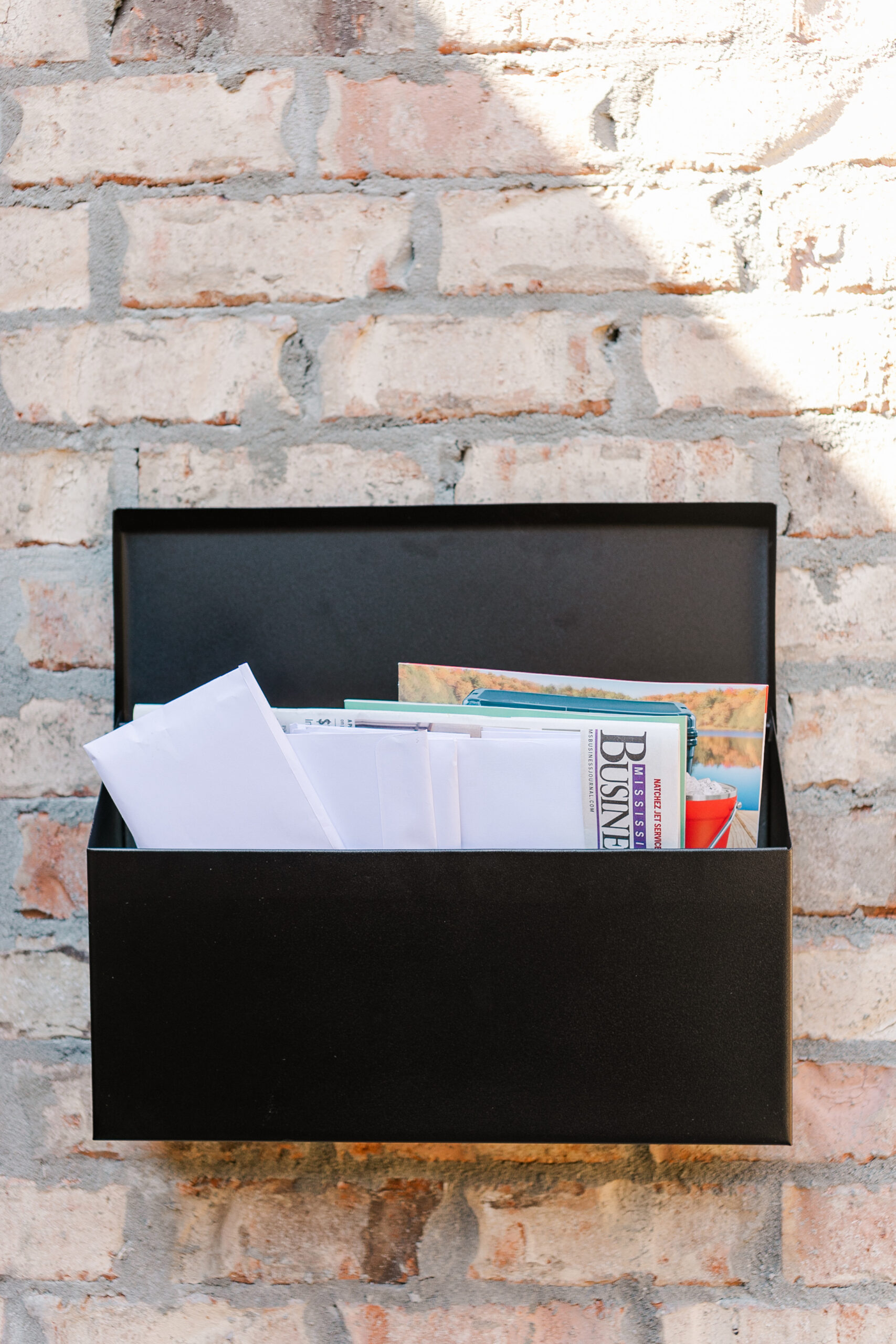 Black wall-mounted mailbox filled with scattered envelopes and a newspaper against a rustic brick wall.