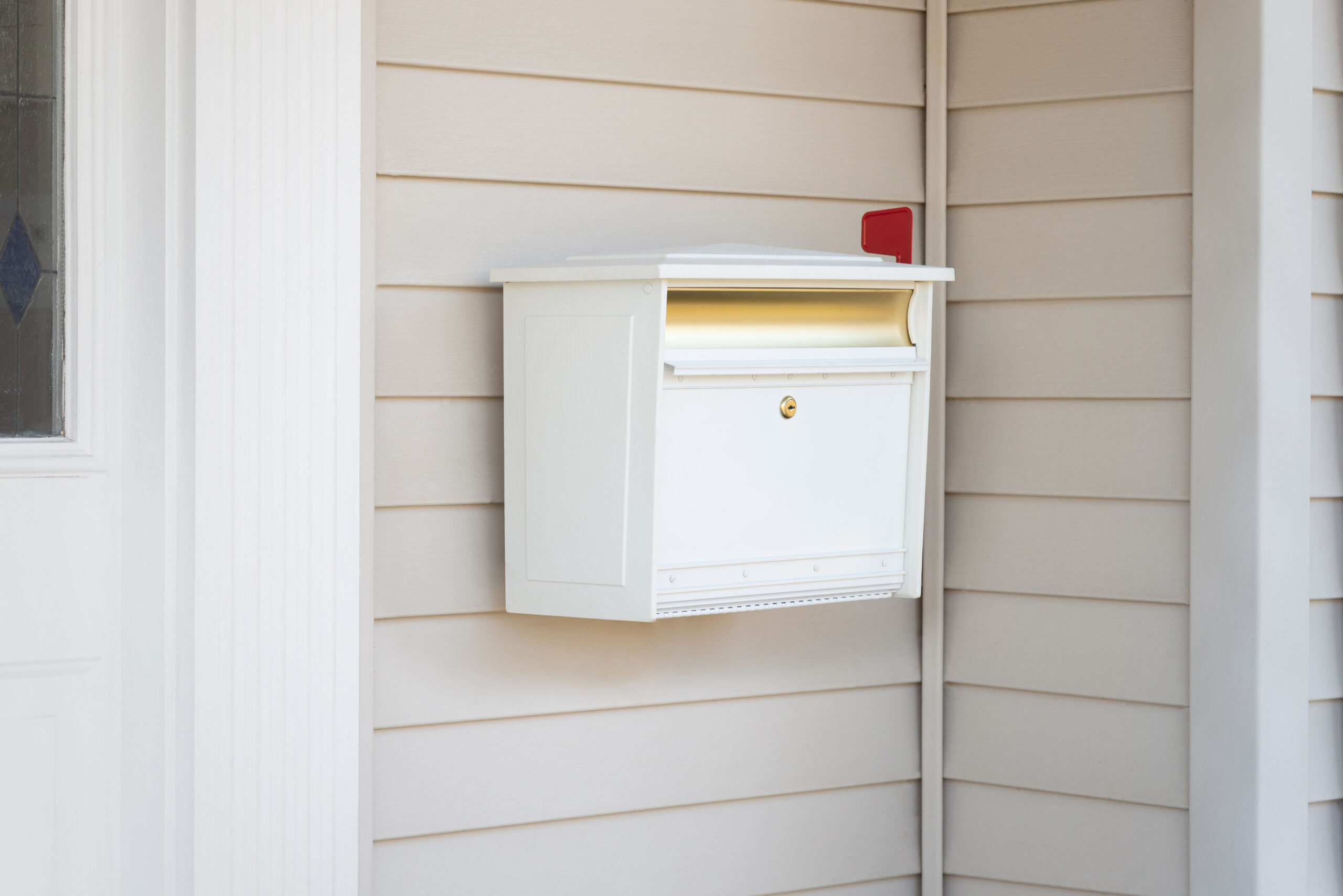 White wall-mounted mailbox on beige siding with a red flag up near a doorway.
