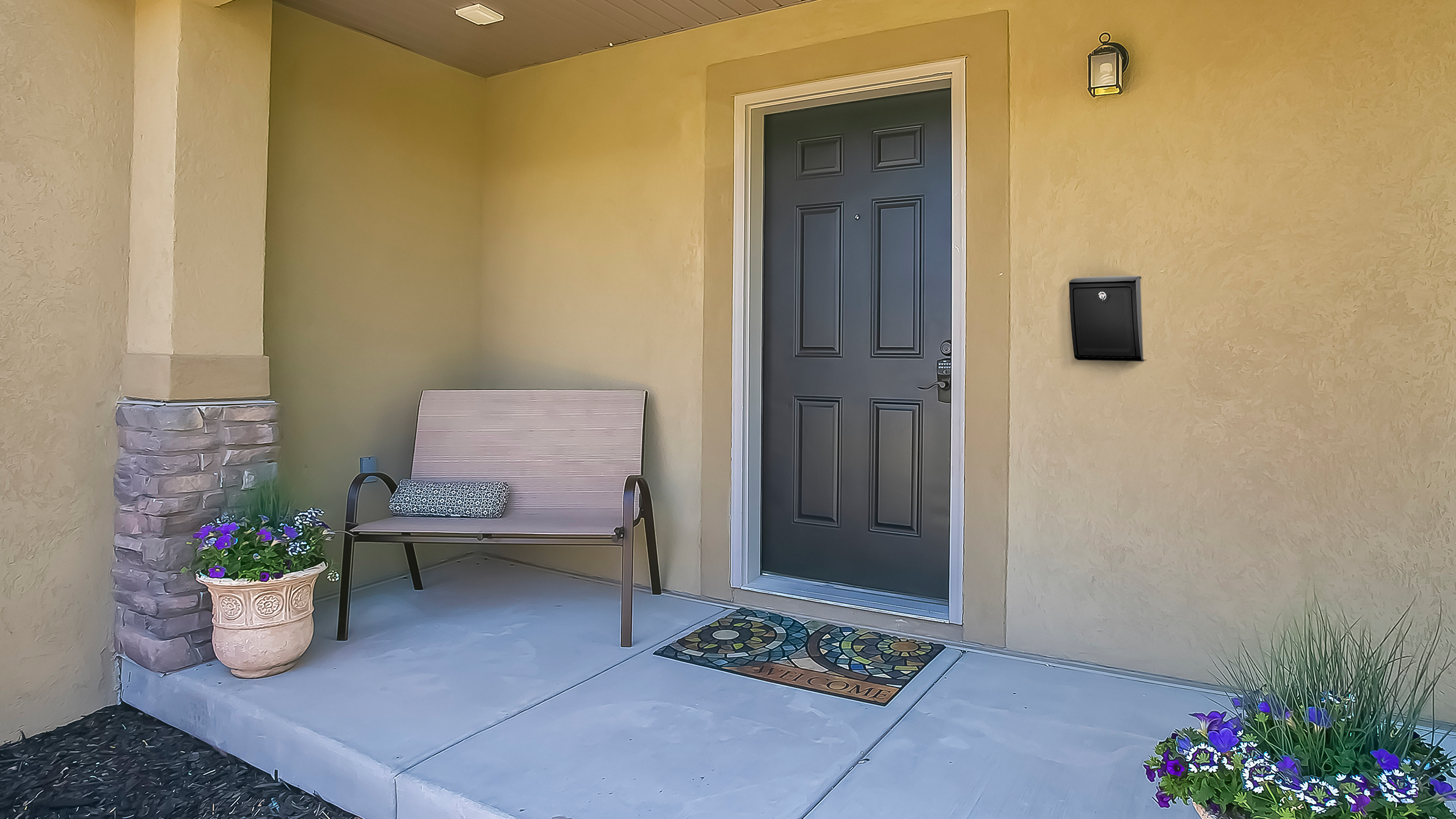 Front porch with a dark gray front door, beige walls, and a metal-framed bench beside a large potted flower plant on the left. A decorative mosaic welcome mat sits on the concrete before the door, with a wall light and mailbox on the right.