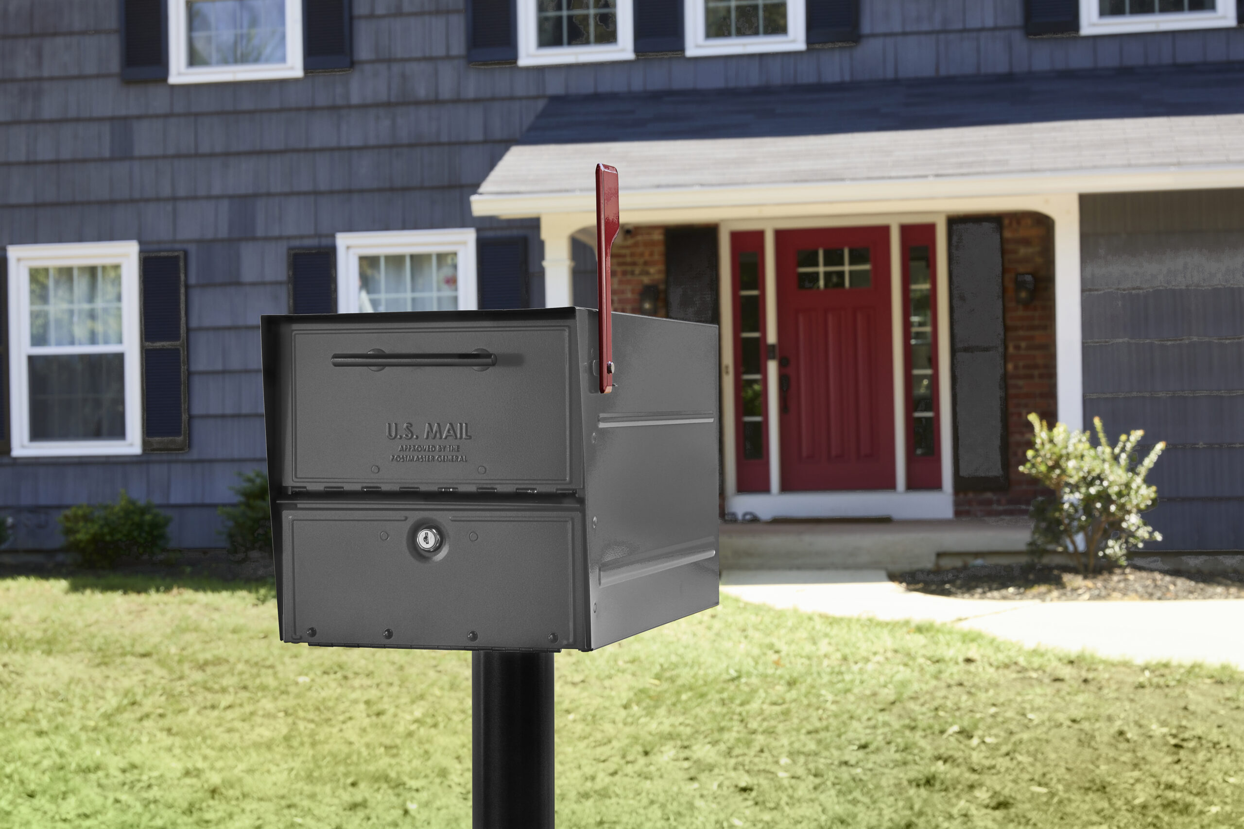 Gray U.S. Mailbox with red flag raised on a black post in front of a blue house with a red front door and green lawn