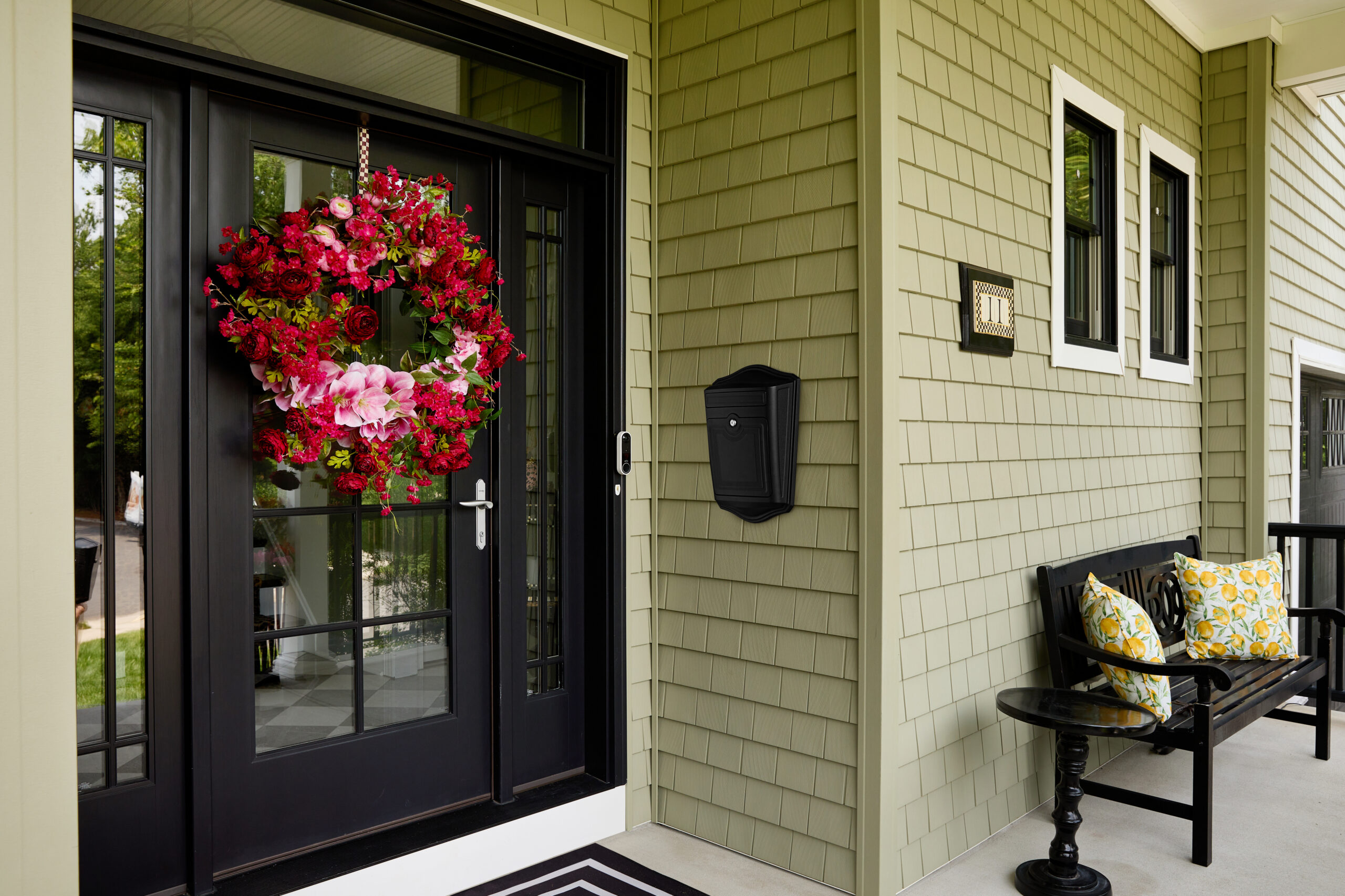 Black front door with a large pink-red floral wreath hanging on it, beige siding, and a striped doormat at the entrance