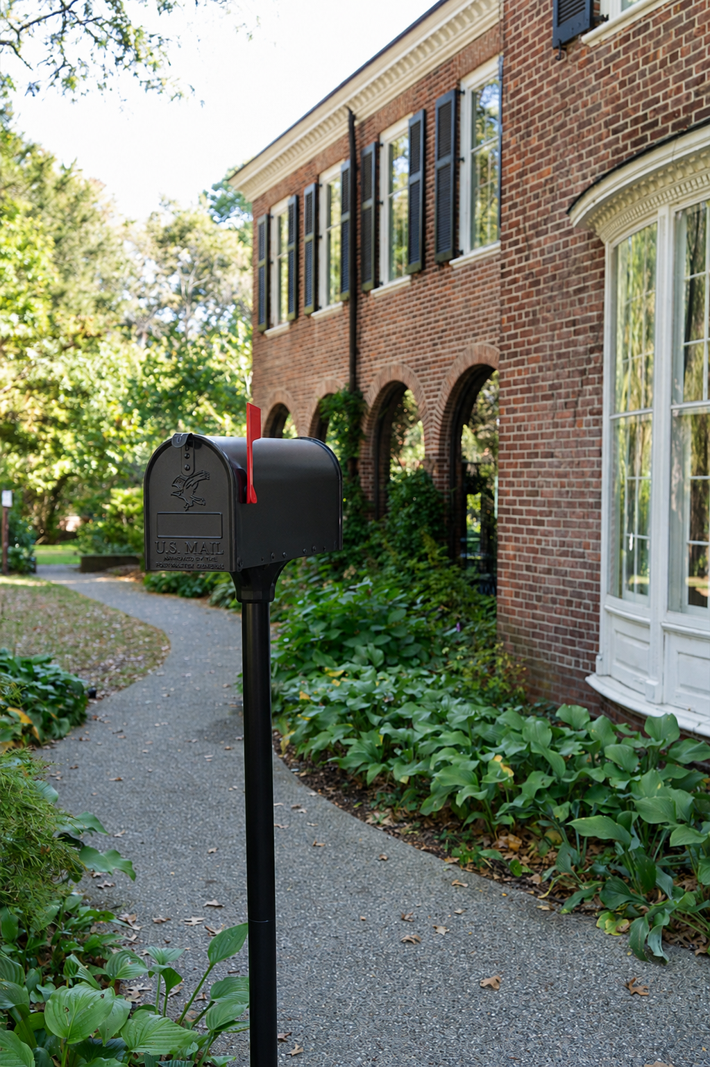 Black U.S. mailbox on a post with a raised red flag beside a brick house with arched windows and lush greenery along a curved path.