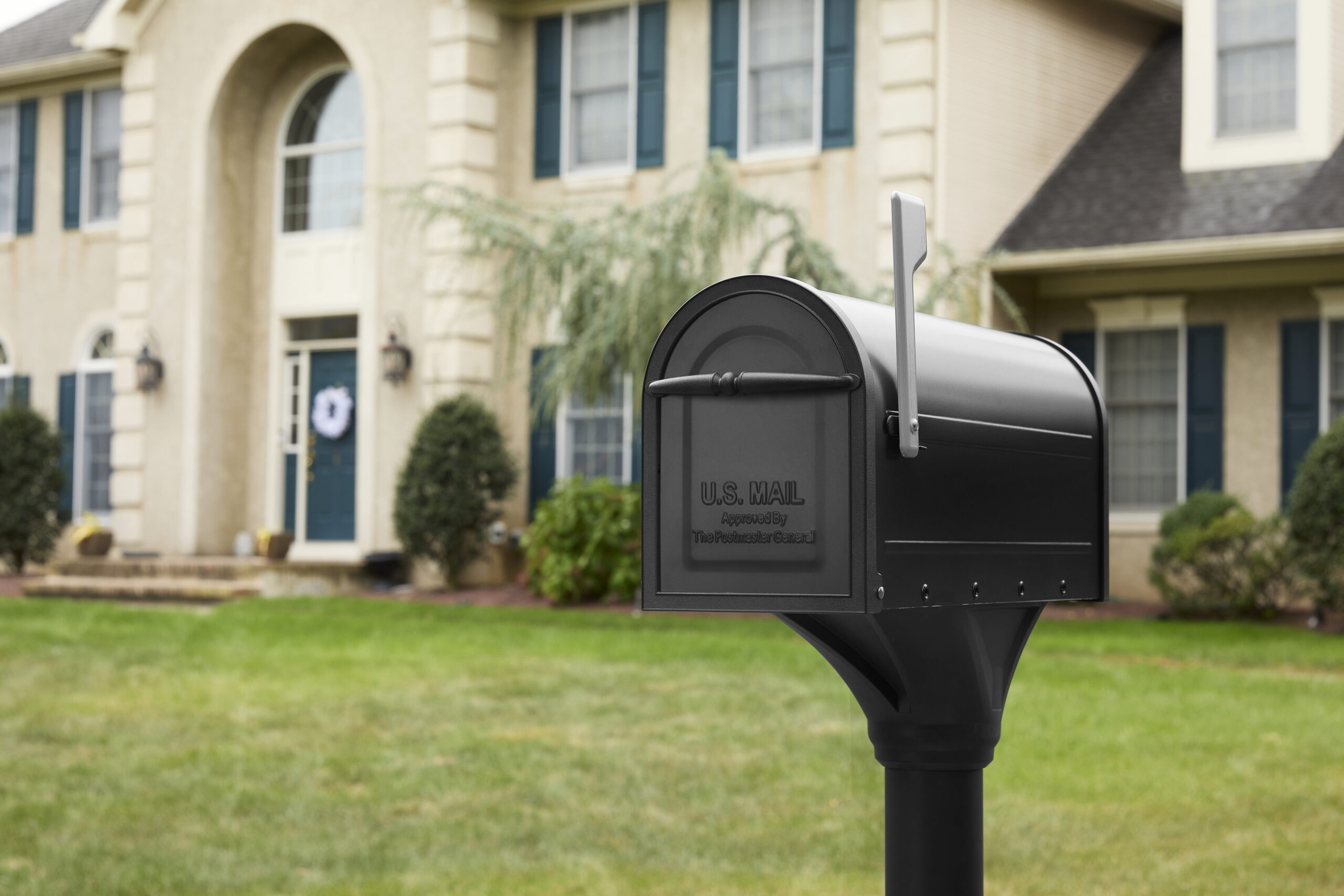 Black U.S. mailbox on a post in a suburban front yard with a green lawn and a large house in the background.