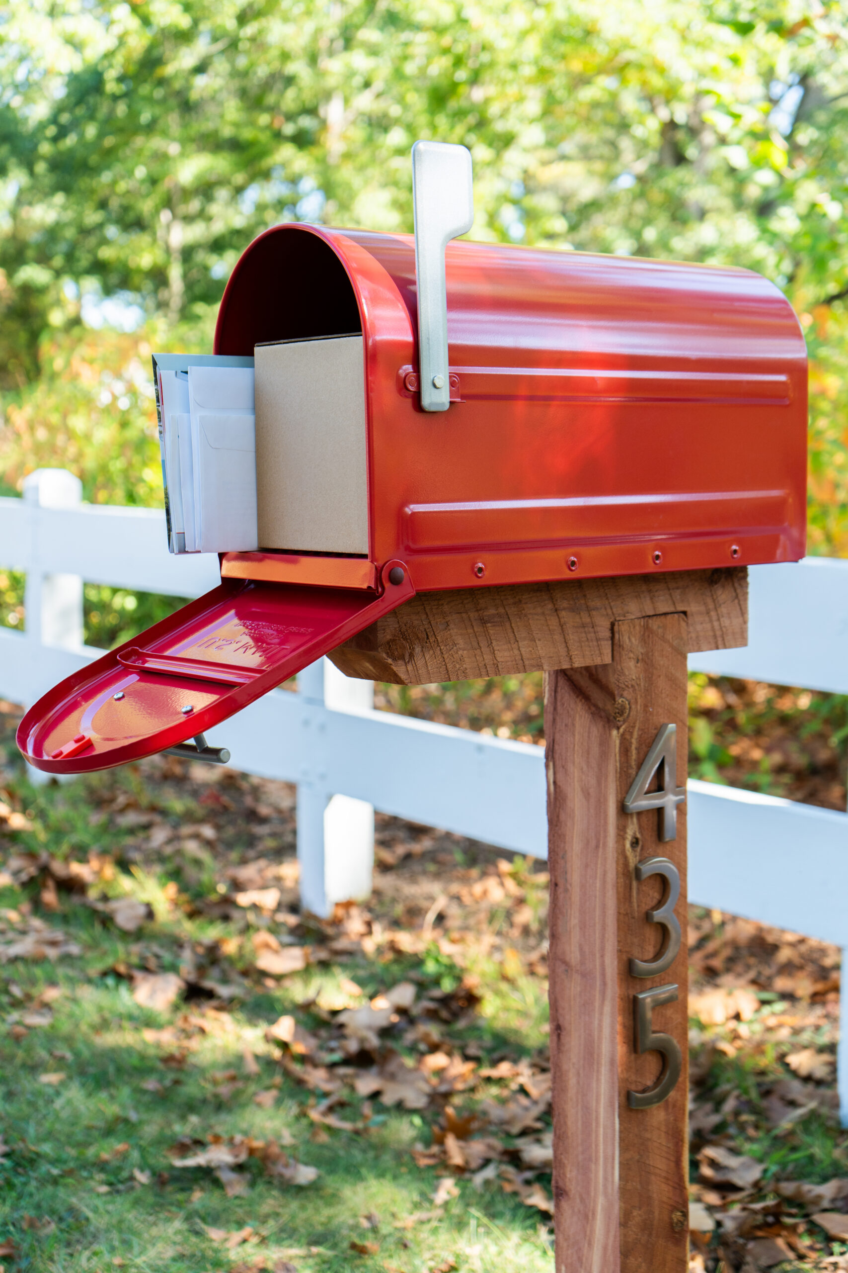 Red mailbox on a wooden post with its door open and mail inside, beside a white fence in a yard.