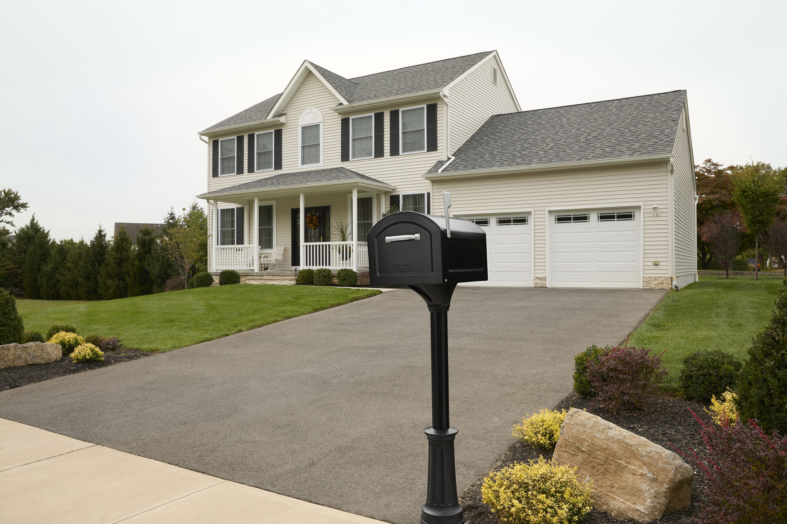 Two-story beige suburban house with white trim, a front porch, and a black mailbox on a post in the driveway.