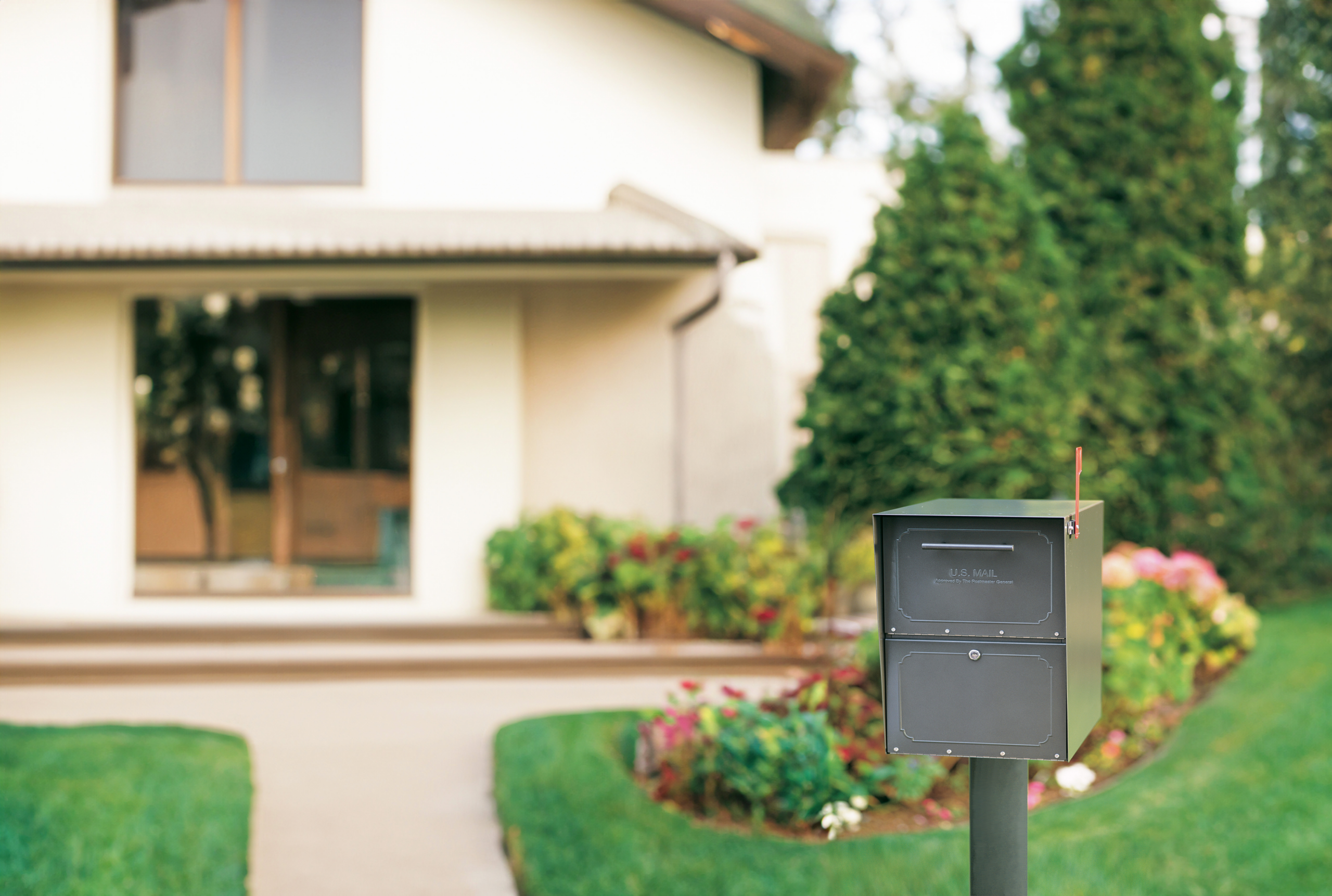 Grey mailbox on a post in the front yard of a suburban home, with a blurred house and landscaping in the background.