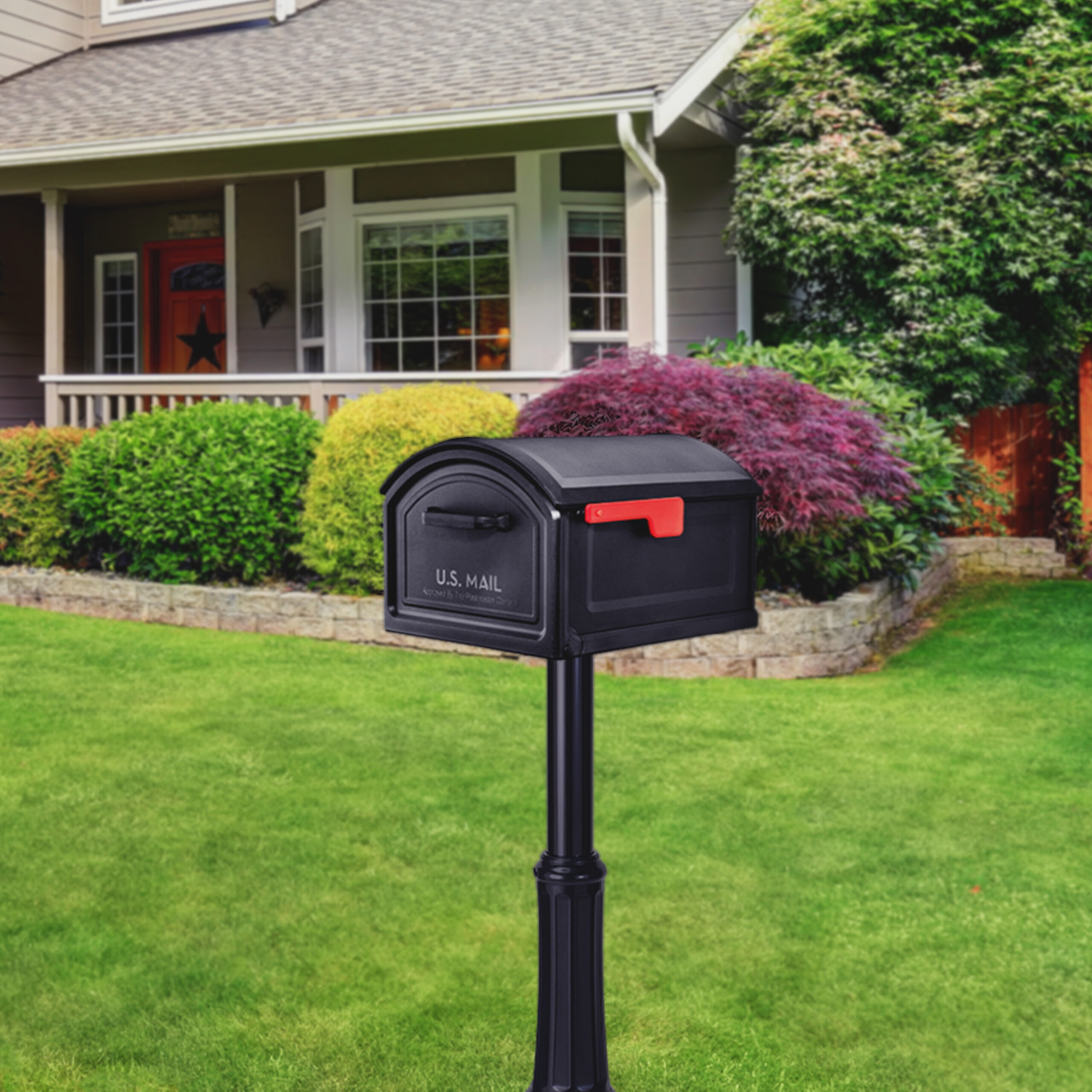 Black U.S. mailbox on a tall post in a front yard with a green lawn and a house in the background.