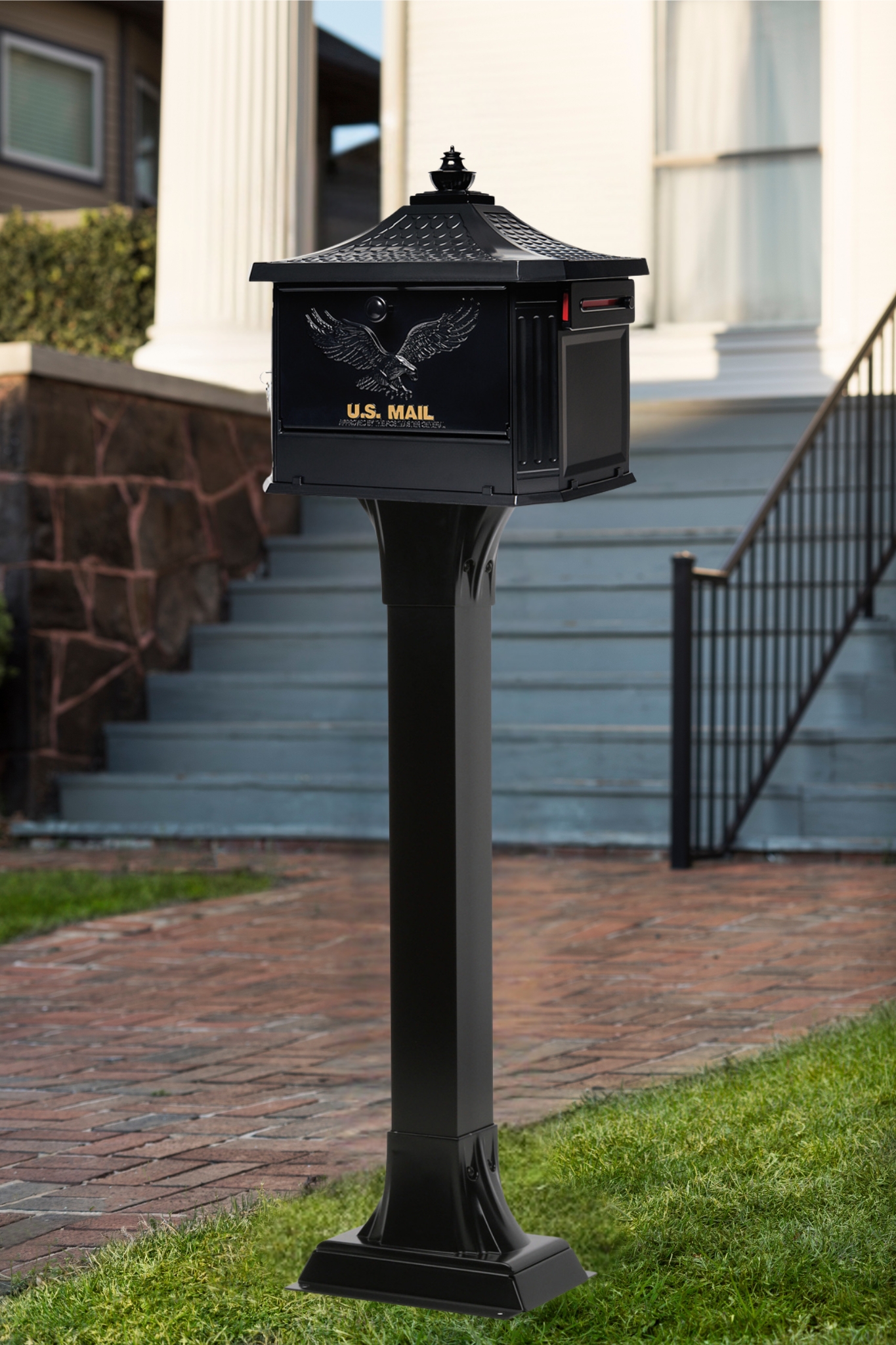 Black US Mail mailbox mounted on a tall post in front of a house with a brick walkway and stairs in the background.