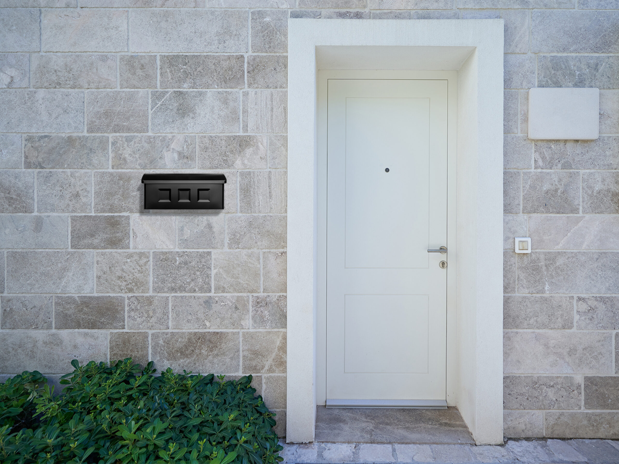 White paneled front door set in a light stone wall, with a black wall mailbox to the left and green shrubs at the base.