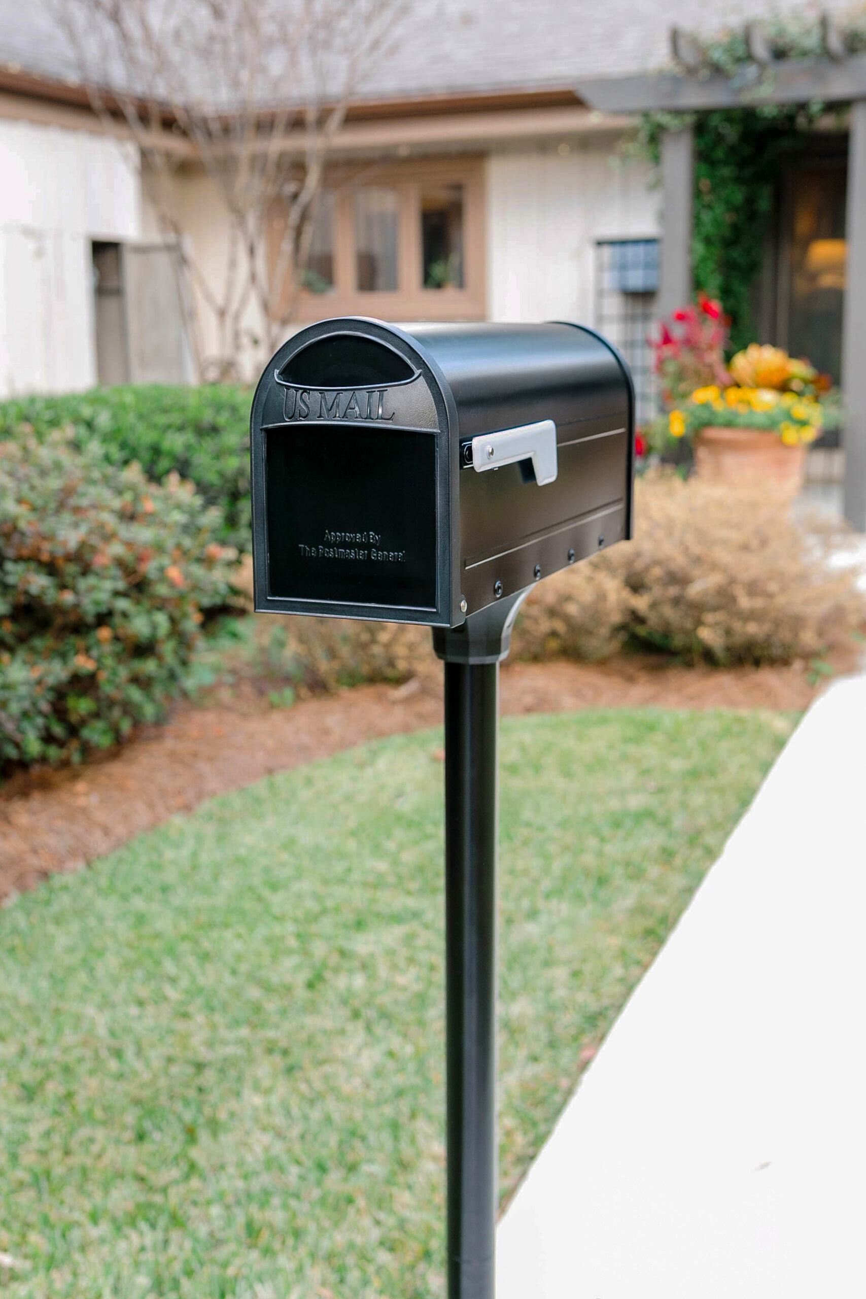 Black curbside USPS mailbox on a metal post in a suburban front yard with a house and flowers in the background