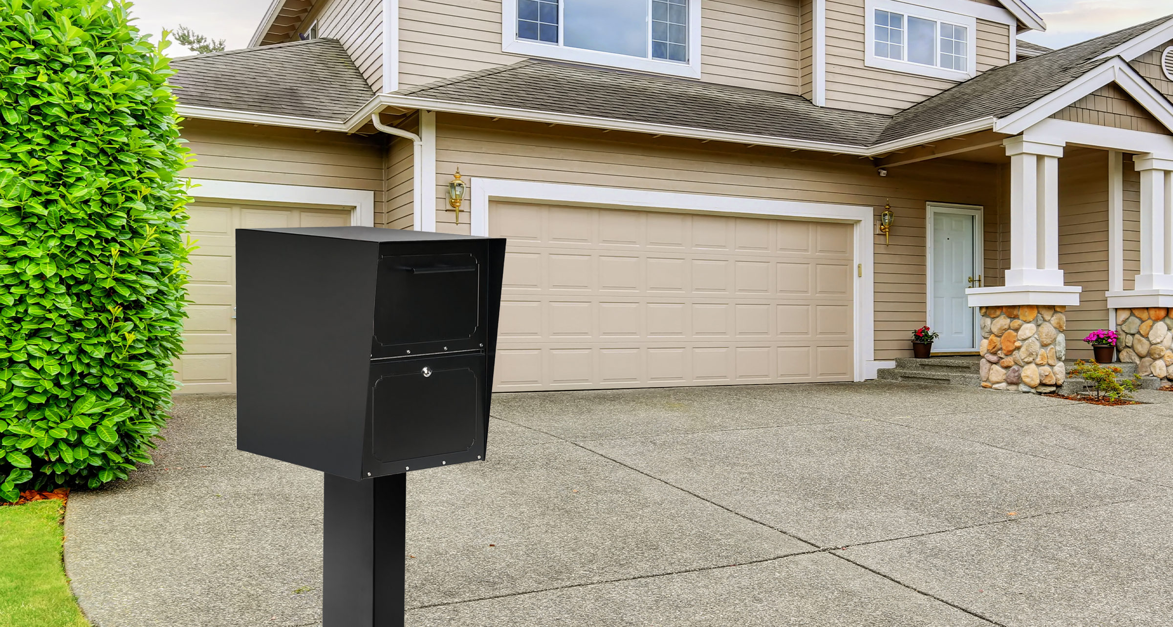 Black metal mailbox on a tall post in a suburban driveway, with a beige two-car garage and a beige siding house in the background.