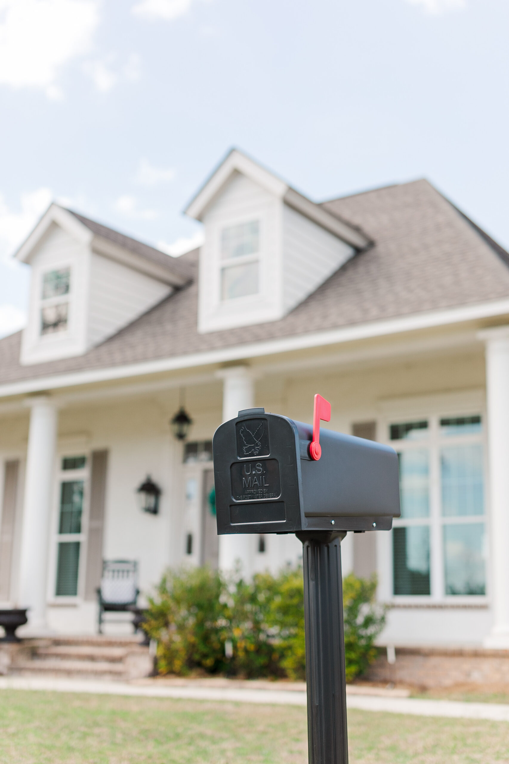 Black USPS mailbox on a tall post in a front yard, with a white suburban house and dormer windows blurred in the background.
