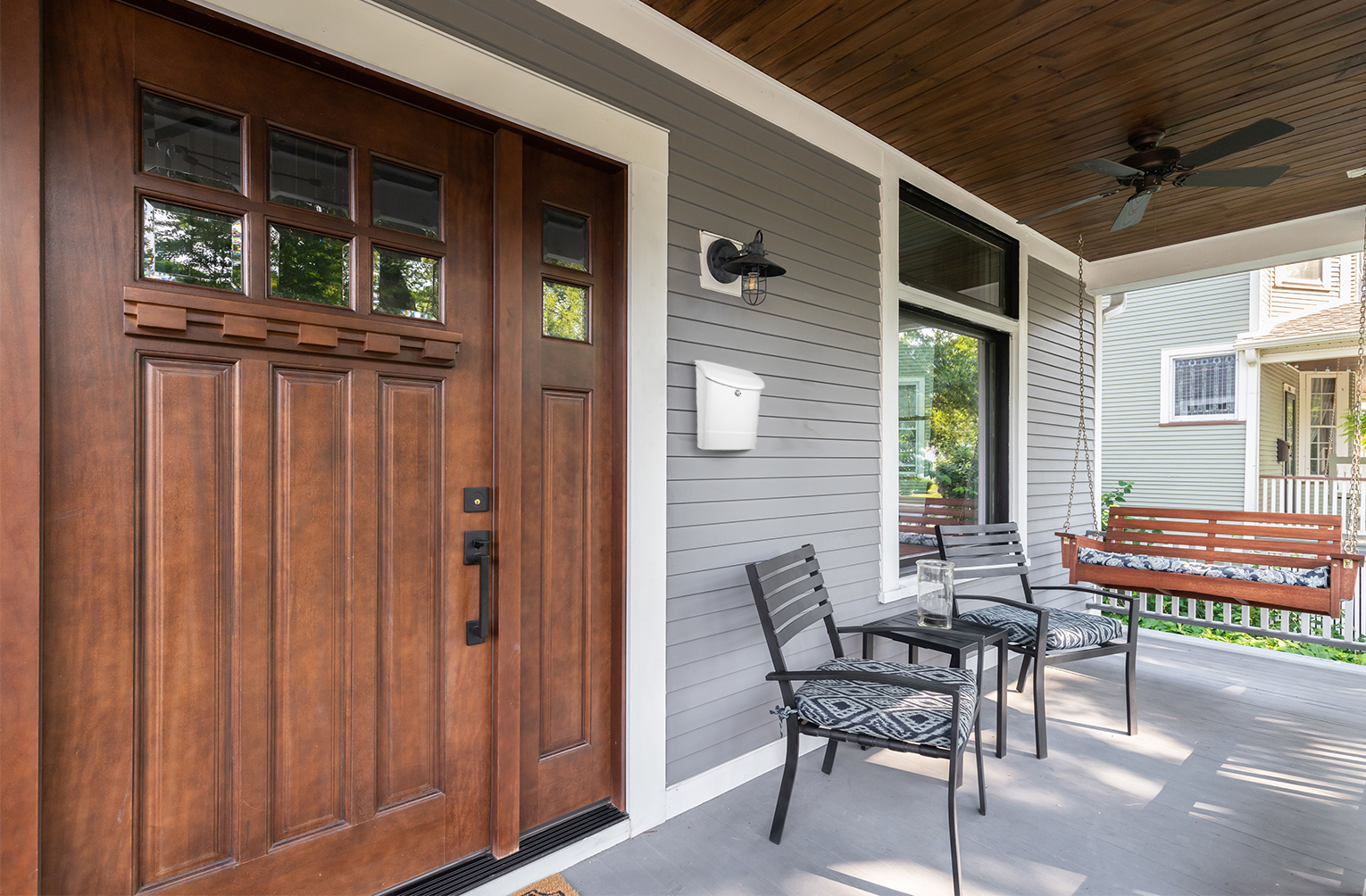 Front porch with a dark wood paneled front door, glass window panes, and a gray-sashed siding wall; two metal chairs and a bench swing on the porch.