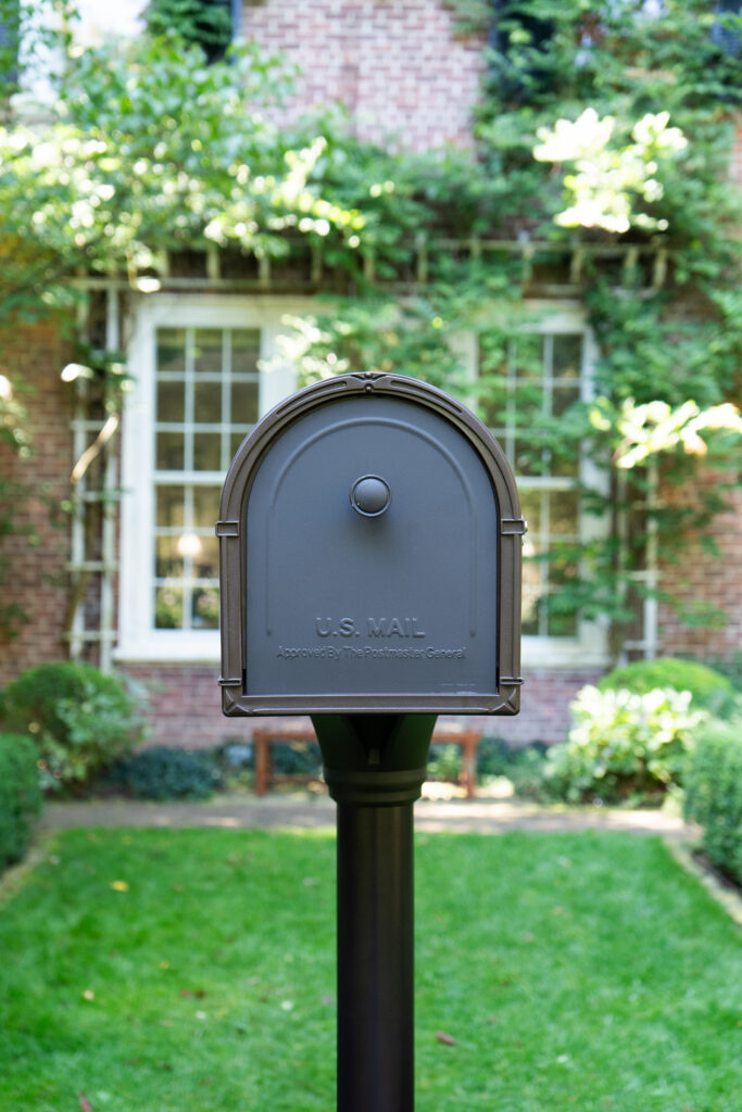 Grey curved-top U.S. Mail mailbox on a post in a suburban front yard, with a brick house and lush greenery in the background.