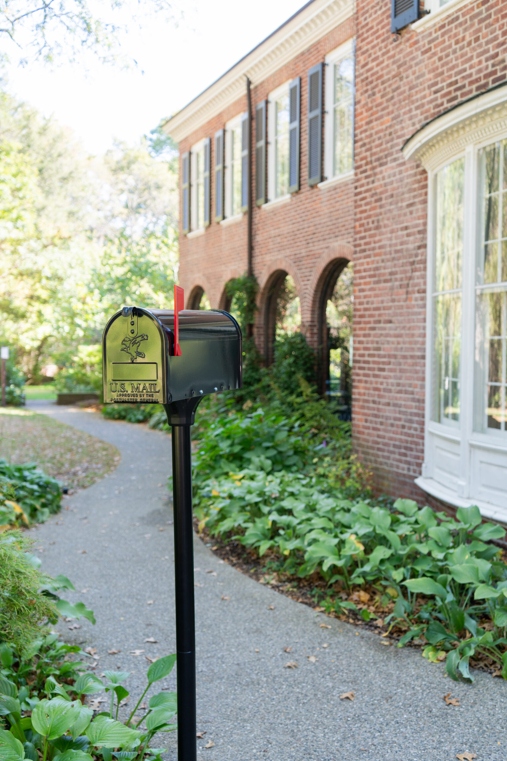 Black U.S. Mail mailbox on a post beside a curved garden path and brick townhouse facade with arched entryways nearby.