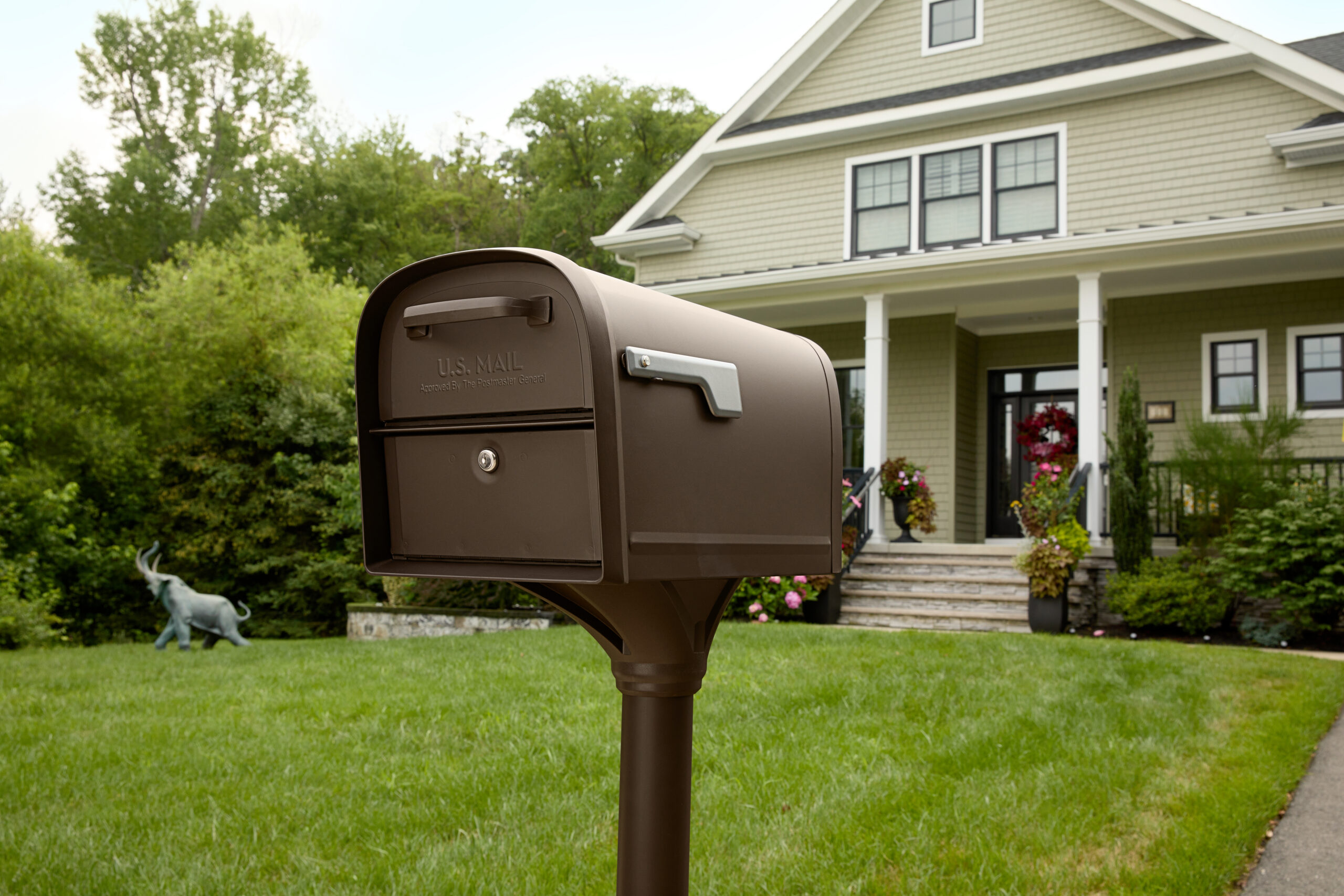 Brown U.S. Mailbox on a post in the front yard of a beige, two-story house with flowers on the steps and a decorative elephant statue in the background.