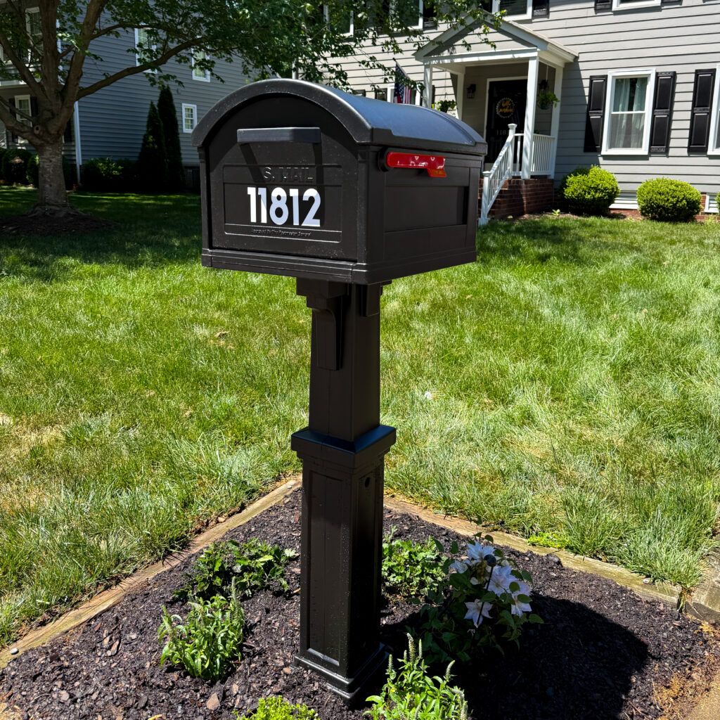 Black mailbox with curved top and bold white house number 1812 in a landscaped front yard. Alternating siding house visible in the background.