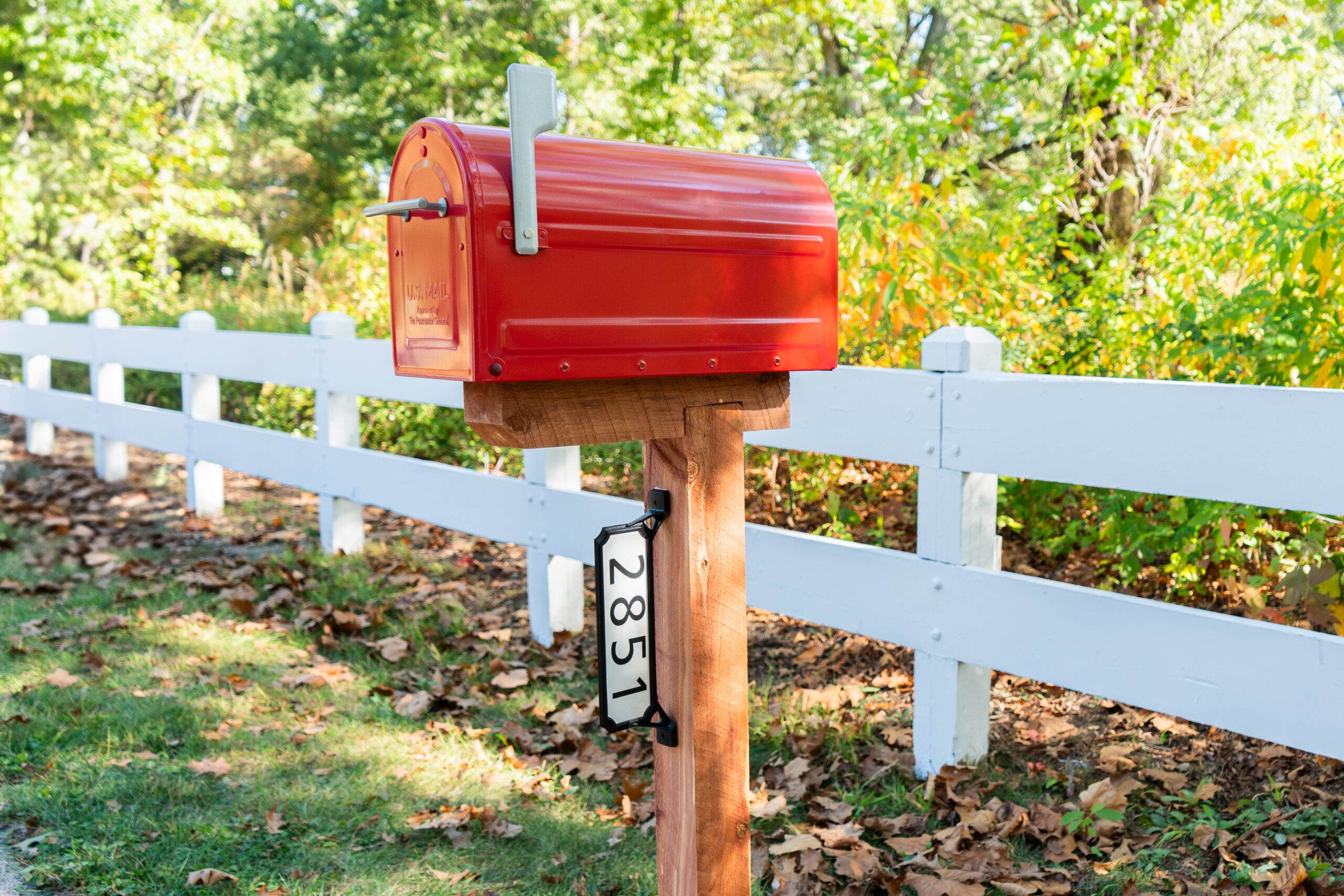 Red curbside mailbox on a wooden post beside a white fence, with fallen leaves and greenery in the background.
