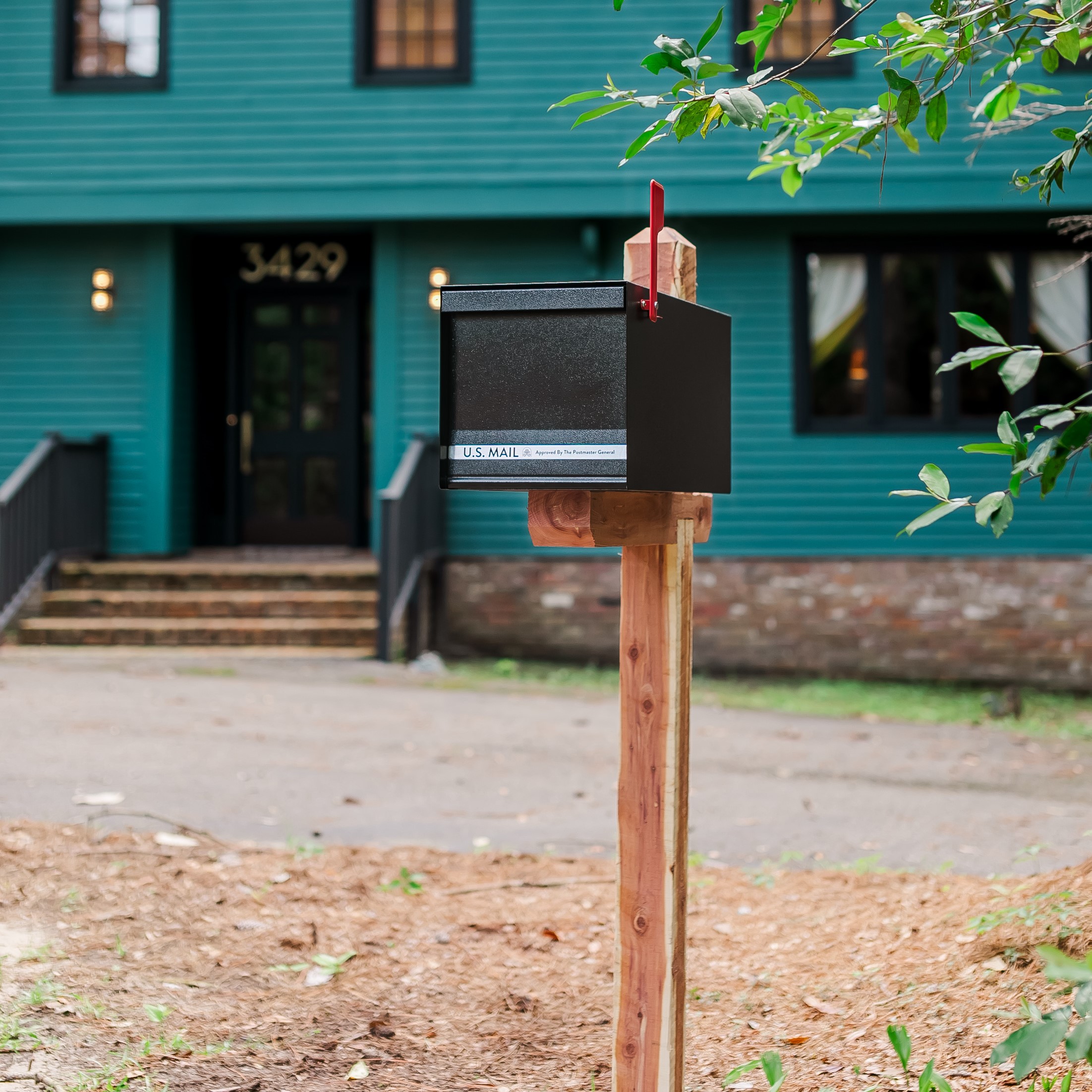 Black mailbox with a red flag on a wooden post in front of a teal house; USPS label visible reading U.S. Mail.