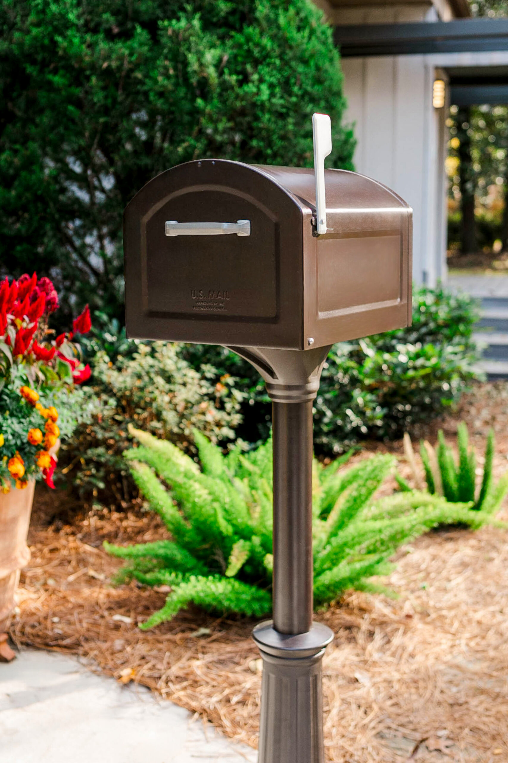 Brown mailbox on a tall post in a landscaped front yard with ferns and bright red flowers nearby.