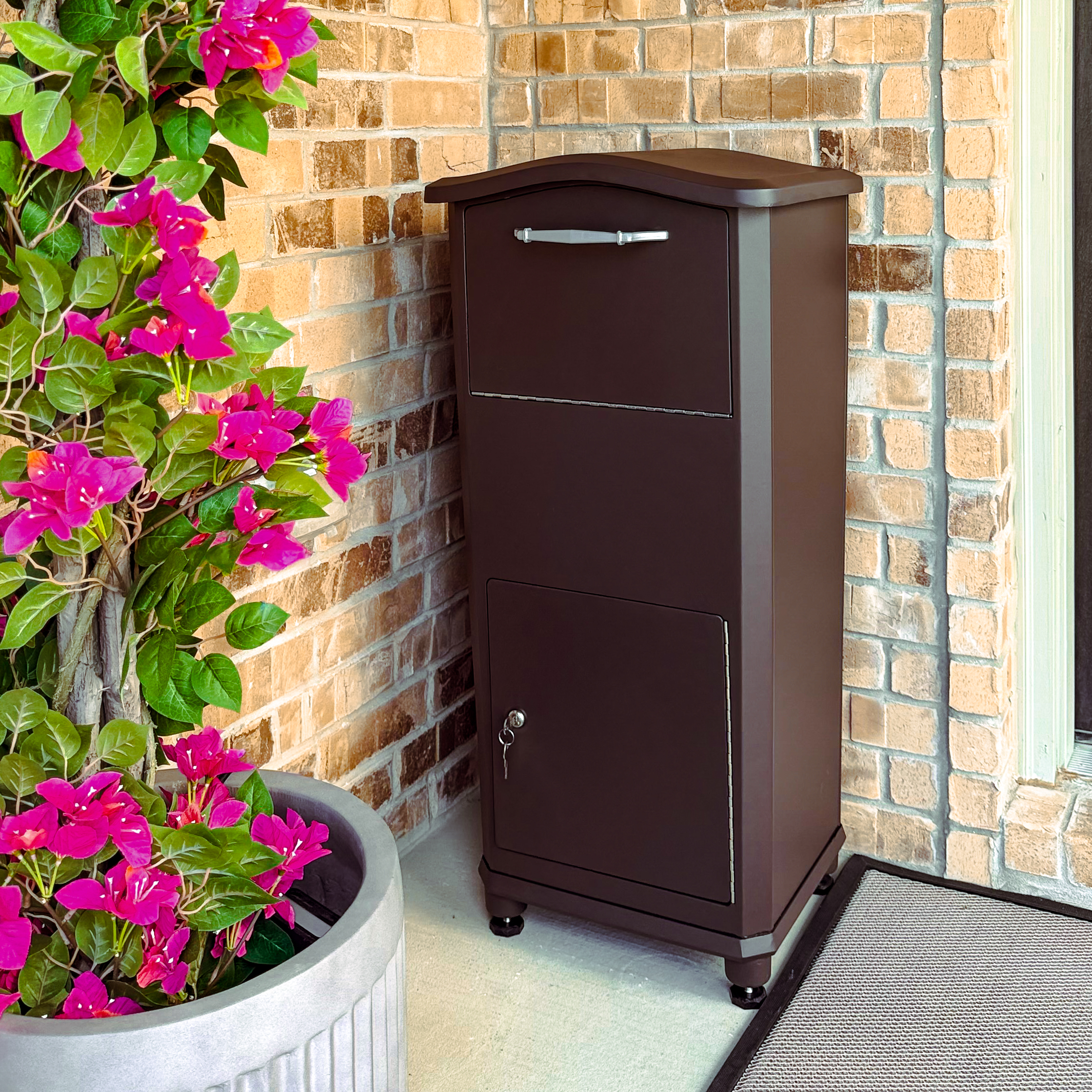 Brown metal mailbox beside a brick wall with vibrant pink flowering plants in a large planter to the left.